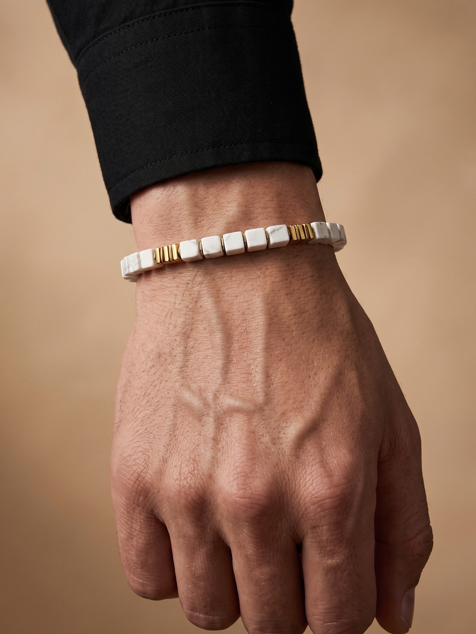 A close-up of a person's hand and wrist wearing a beaded bracelet made of white square beads with small gold accent beads, set against a warm beige background.