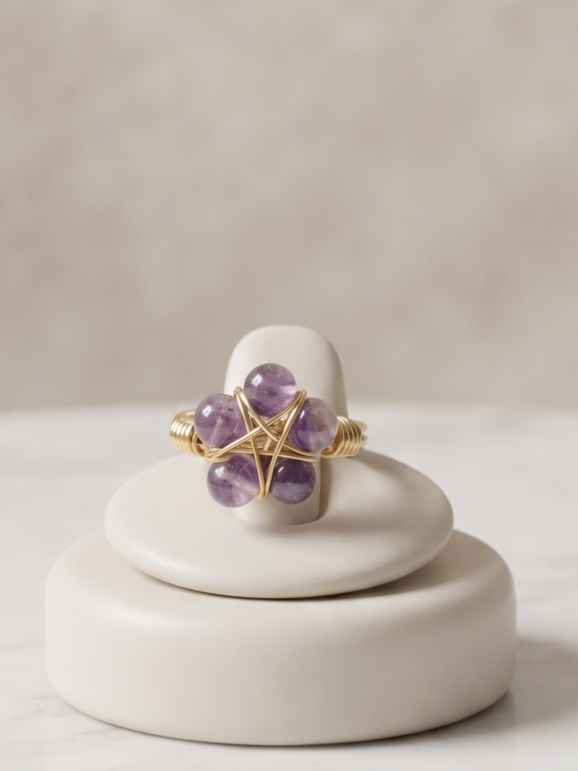 A gold ring with five purple amethyst gemstones arranged in a flower shape, displayed on a small round cream ceramic stand on a white marble surface.