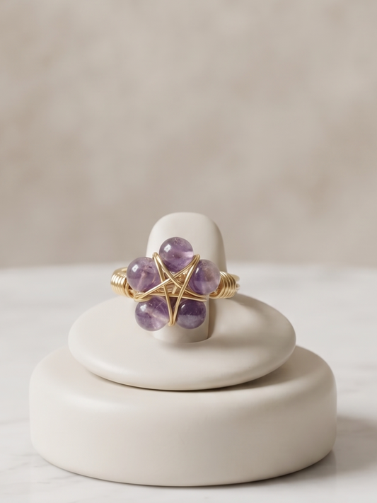 A gold ring with five purple amethyst gemstones arranged in a flower shape, displayed on a small round cream ceramic stand on a white marble surface.