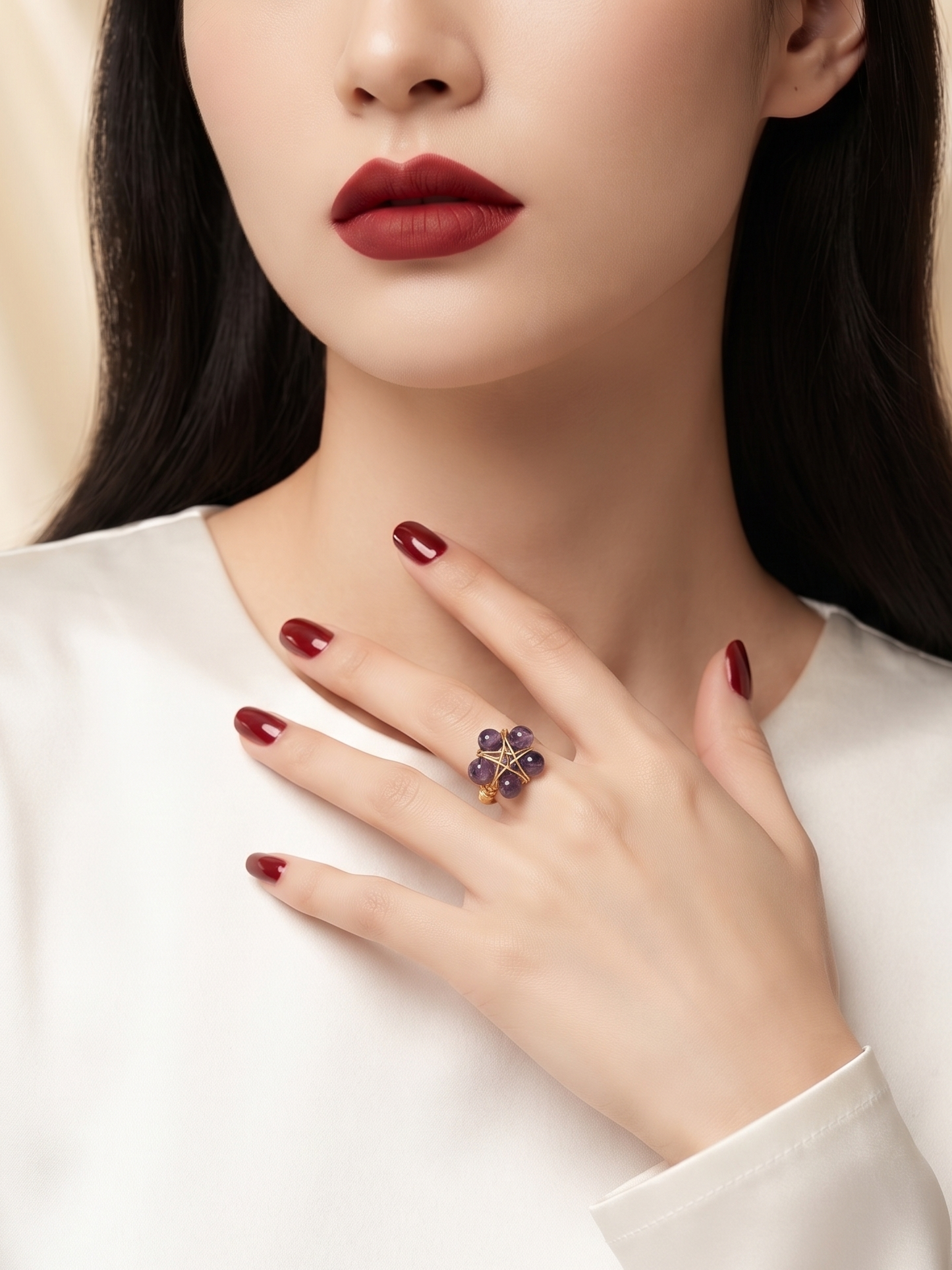 A close-up of a woman wearing a distinctive ring with four dark blue gemstones on her ring finger, styled with dark red nail polish and a white satin top.