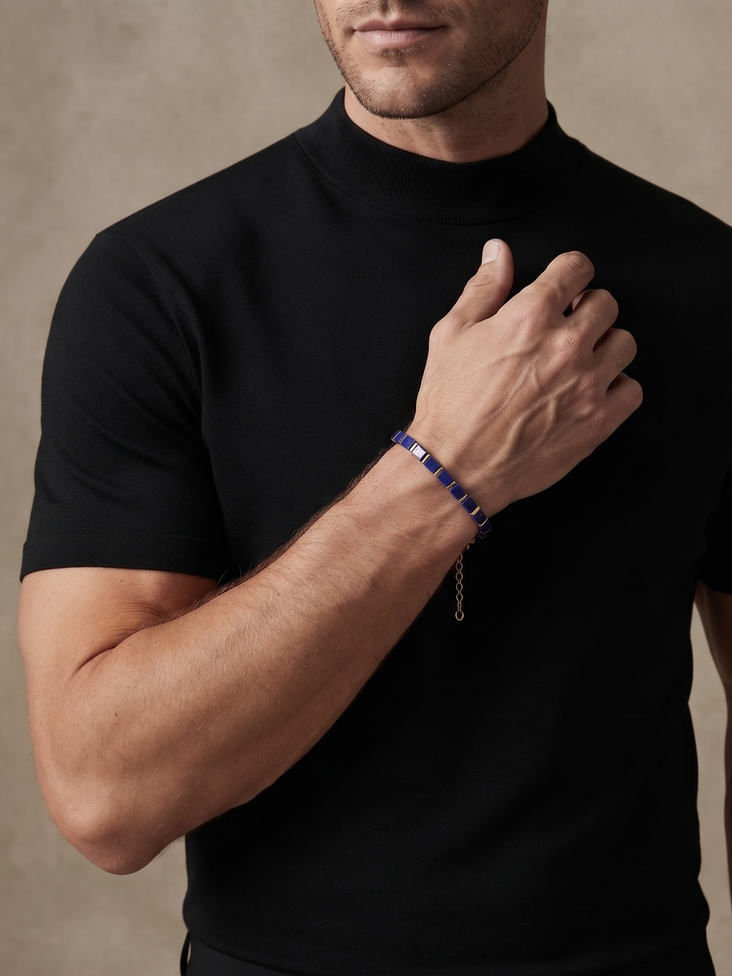 A man wearing a black short-sleeve mock neck t-shirt and black trousers, wearing a dark blue beaded bracelet with a gold chain dangle on his wrist, posed against a textured beige background.
