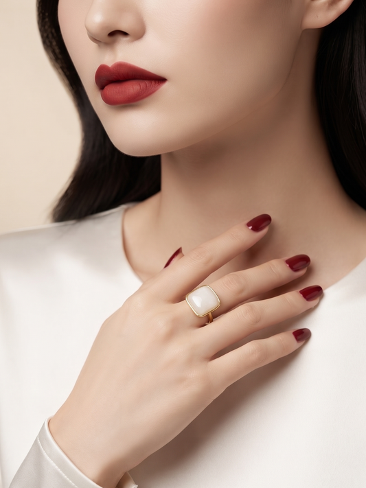 A close-up lifestyle shot of a model wearing a large white oval ring with a gold band, styled with dark red lipstick and manicured nails on a white satin top.