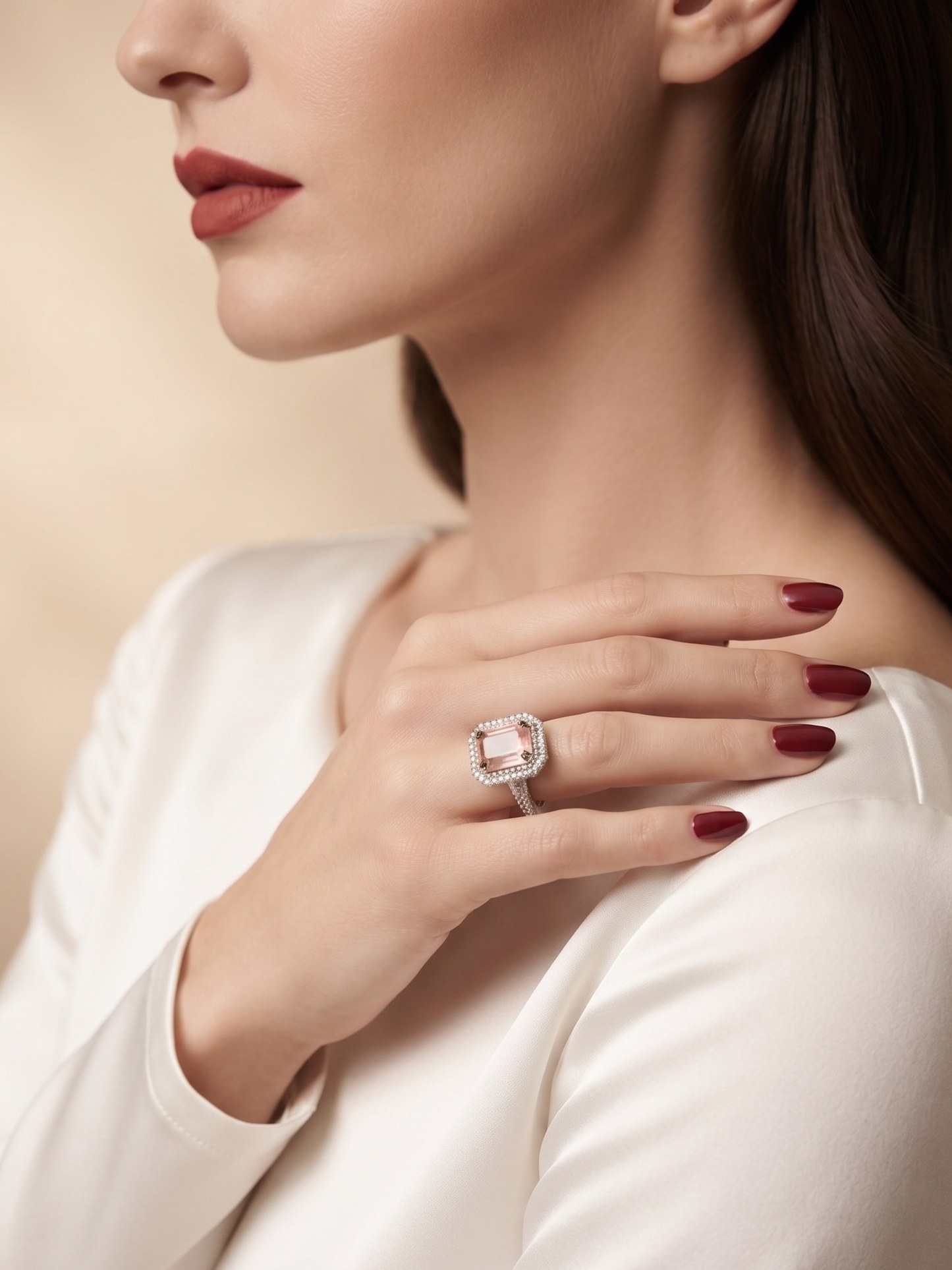 A close-up lifestyle shot of a woman wearing a large cushion-cut pink sapphire ring on her finger, dressed in a white satin top against a warm beige background.