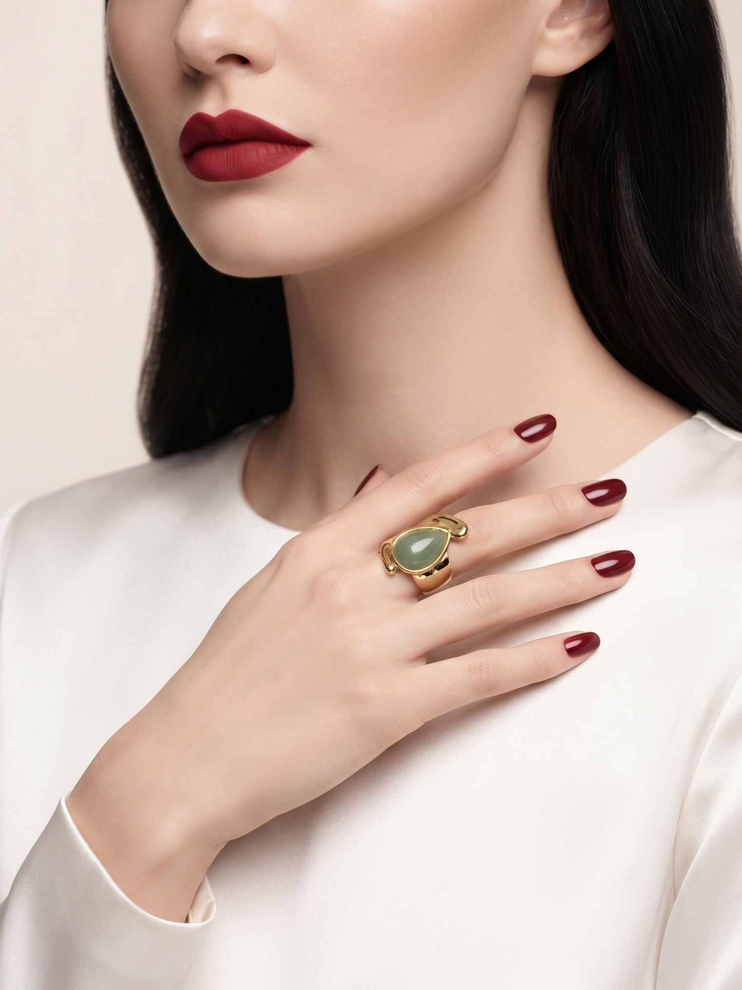 A close-up of a woman wearing a large gold ring with a green oval stone, styled with dark red lipstick and manicured nails, wearing a white satin top.