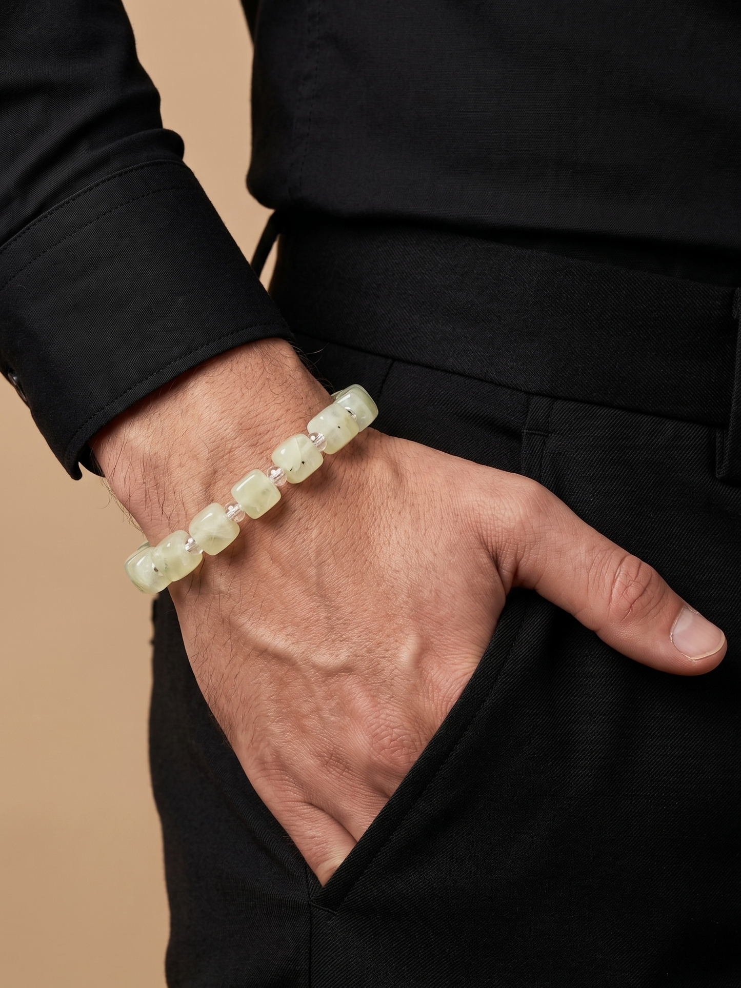 A close-up of a person's hand and wrist wearing a light green jade beaded bracelet, dressed in a black shirt and black trousers against a warm beige background.