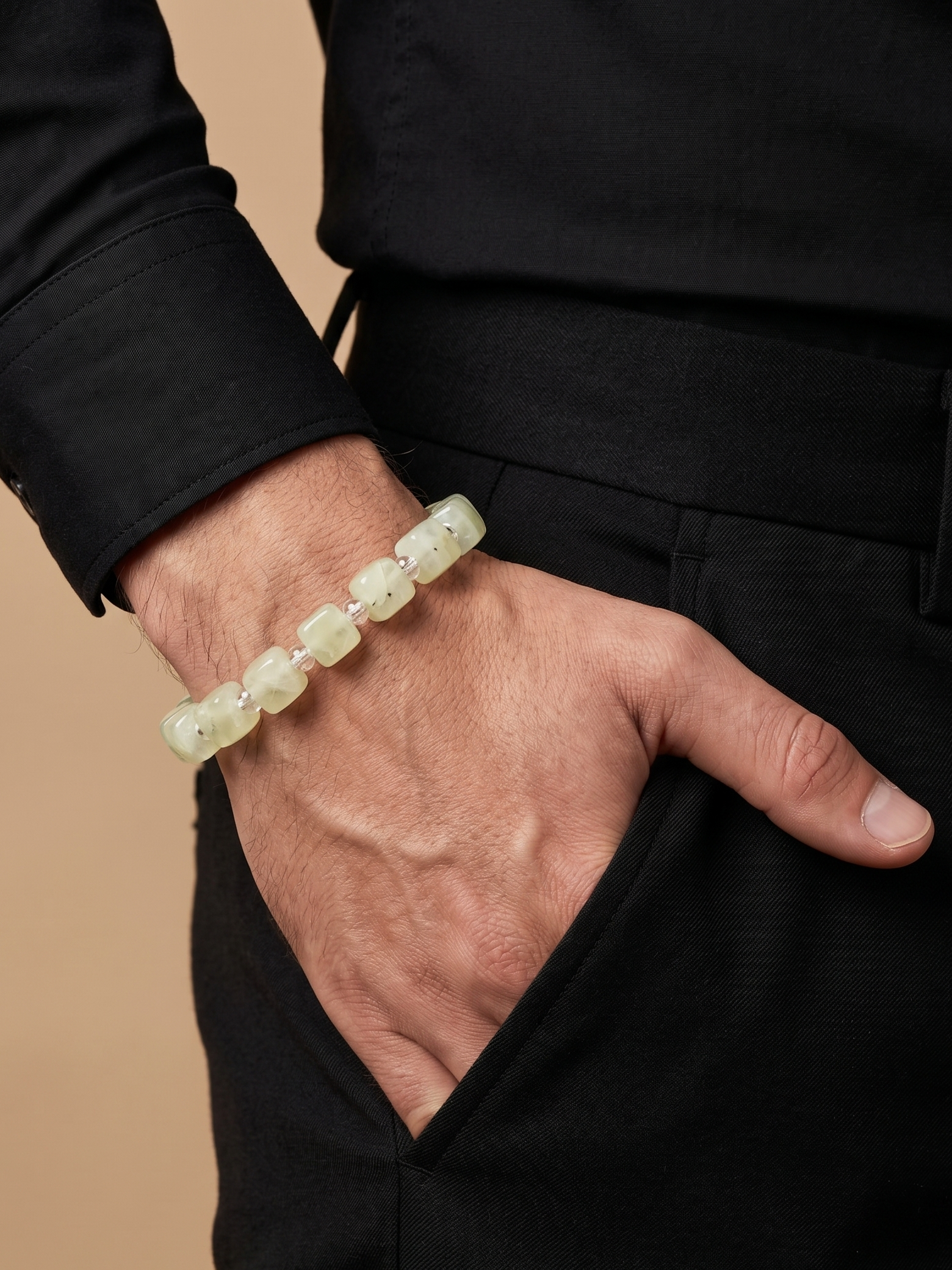 A close-up of a person's hand and wrist wearing a light green jade beaded bracelet, dressed in a black shirt and black trousers against a warm beige background.