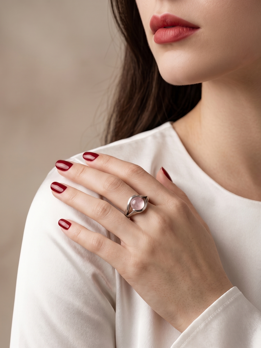 A close-up lifestyle shot of a woman wearing a silver ring with a pink cabochon gemstone on her finger, resting her hand on her shoulder. She is wearing a white long-sleeve top and has dark red nail polish against a soft, neutral beige background.
