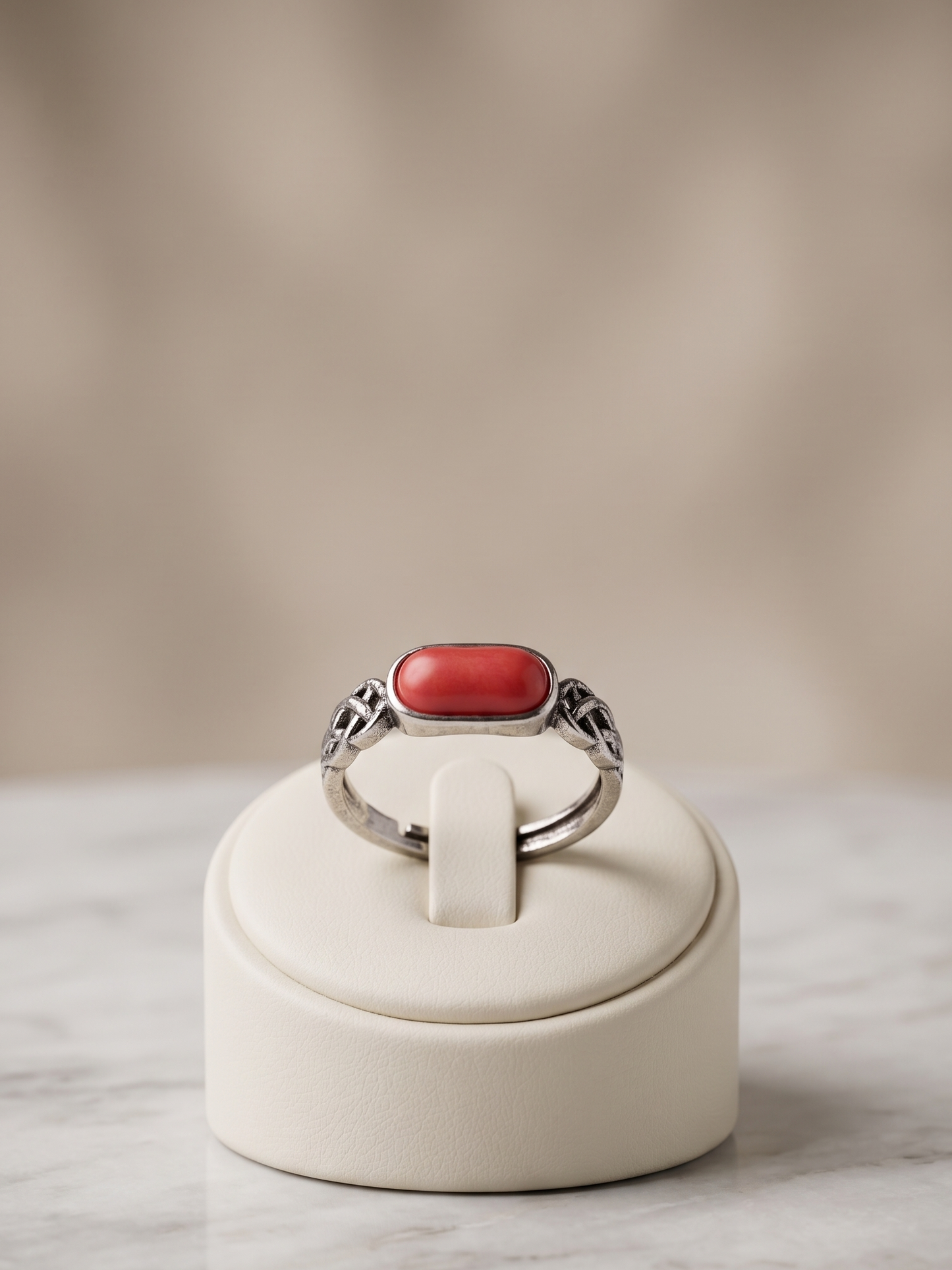 A silver ring with a rectangular red cabochon stone, displayed on a cream-colored jewelry stand over a marble surface with a soft beige background.