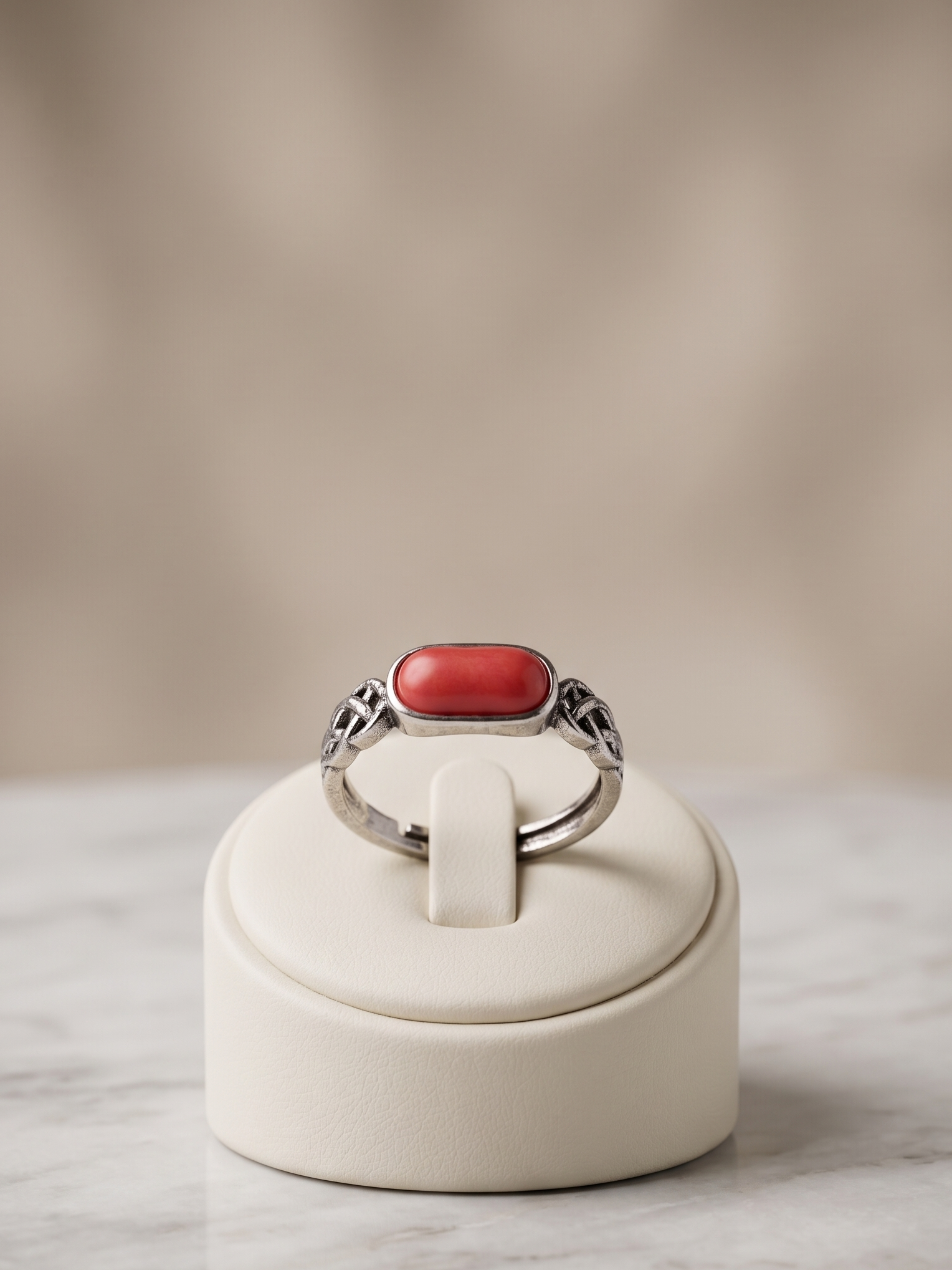 A silver ring with a rectangular red cabochon stone, displayed on a cream-colored jewelry stand over a marble surface with a soft beige background.