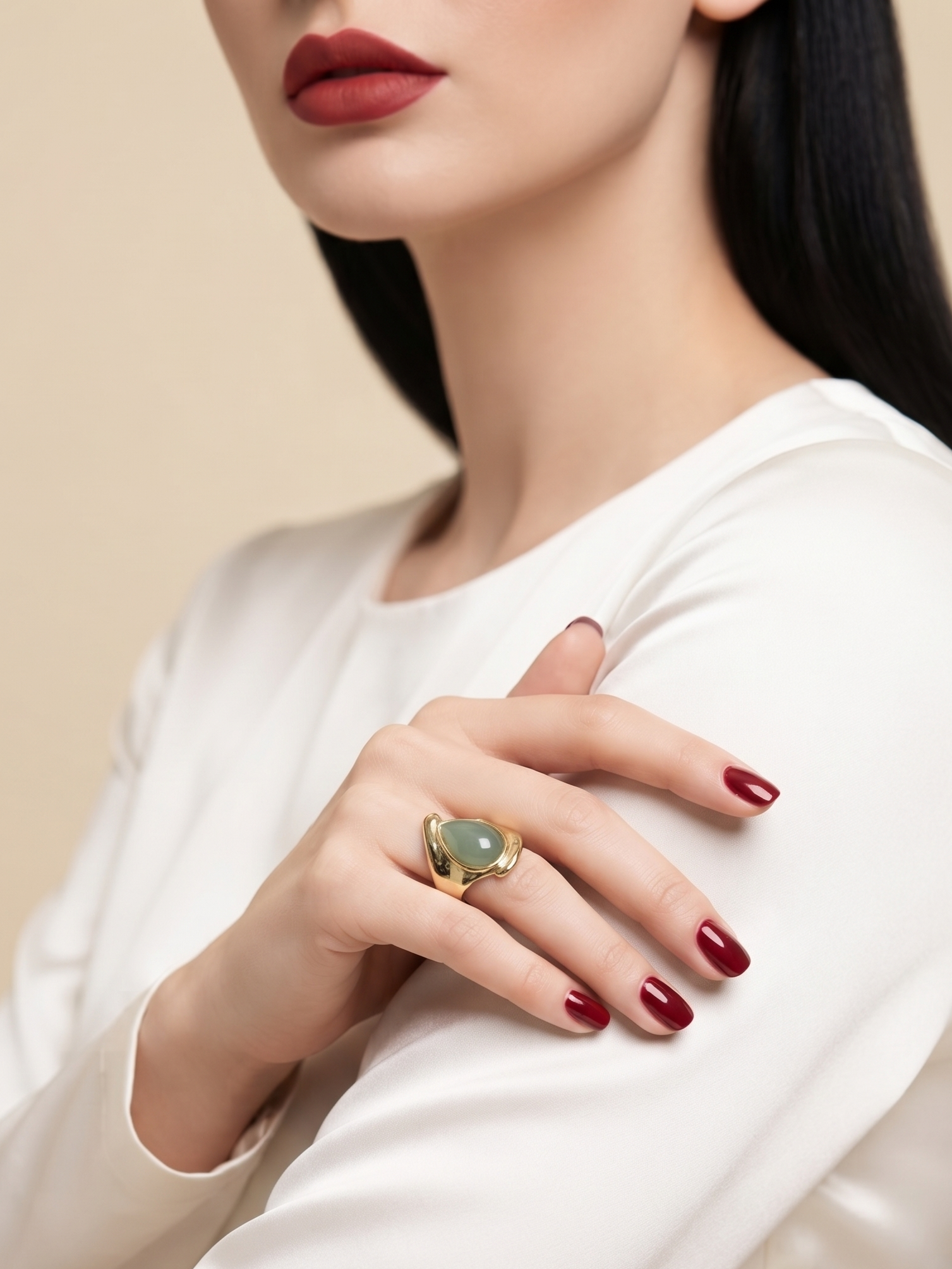 A close-up of a woman wearing a gold ring with a heart-shaped green stone, styled with a white satin top and dark red lipstick.