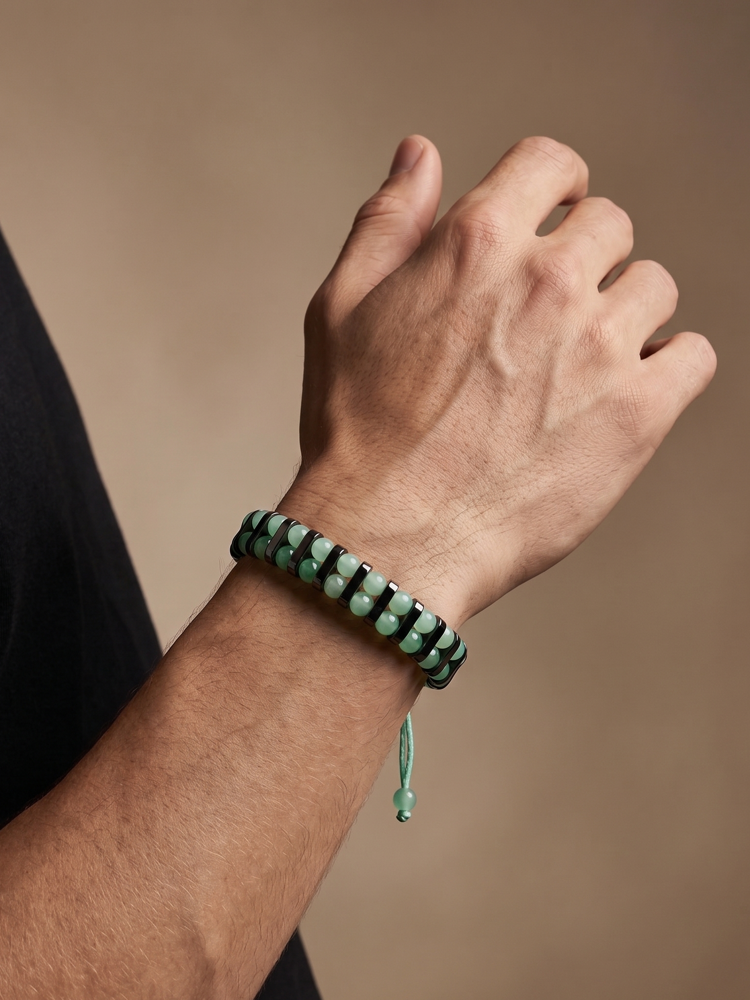 A close-up of a person's hand and wrist wearing a beaded bracelet made of dark green and light green beads with a matching green cord tassel, set against a warm brown background.