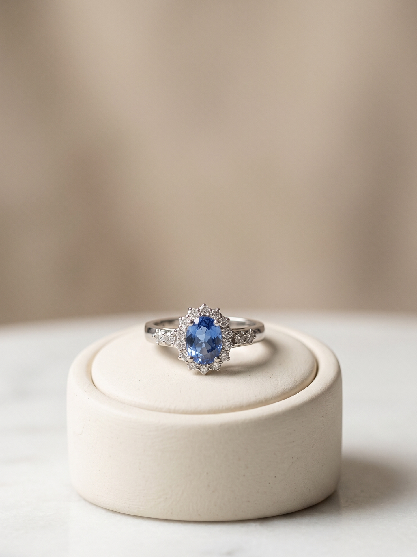 A silver ring with a round blue gemstone and small clear accent stones, displayed on a round cream jewelry stand against a soft beige background.