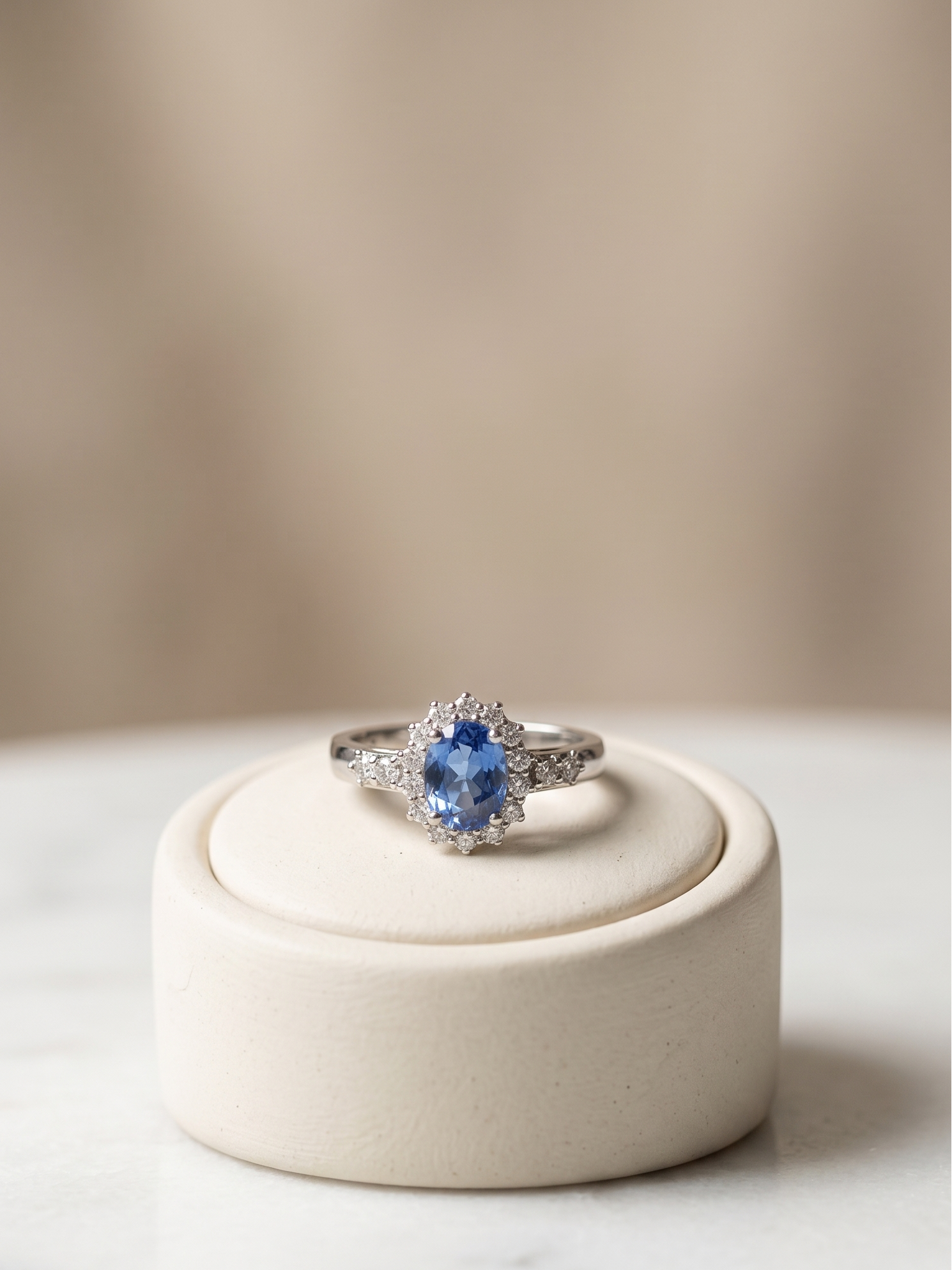 A silver ring with a round blue gemstone and small clear accent stones, displayed on a round cream jewelry stand against a soft beige background.