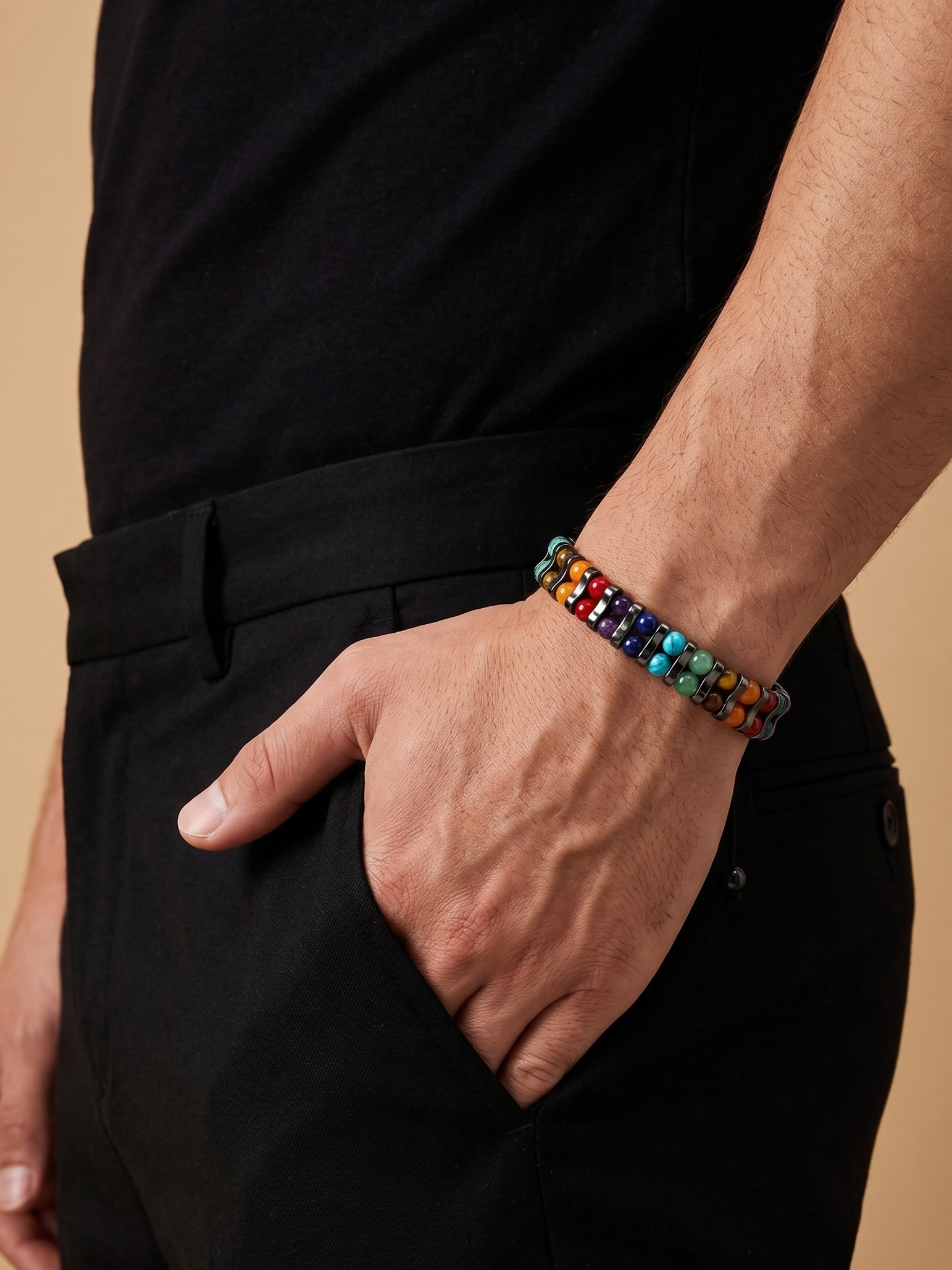 A close-up of a person wearing a colorful beaded bracelet on their wrist, dressed in a black shirt and black trousers against a warm beige background.