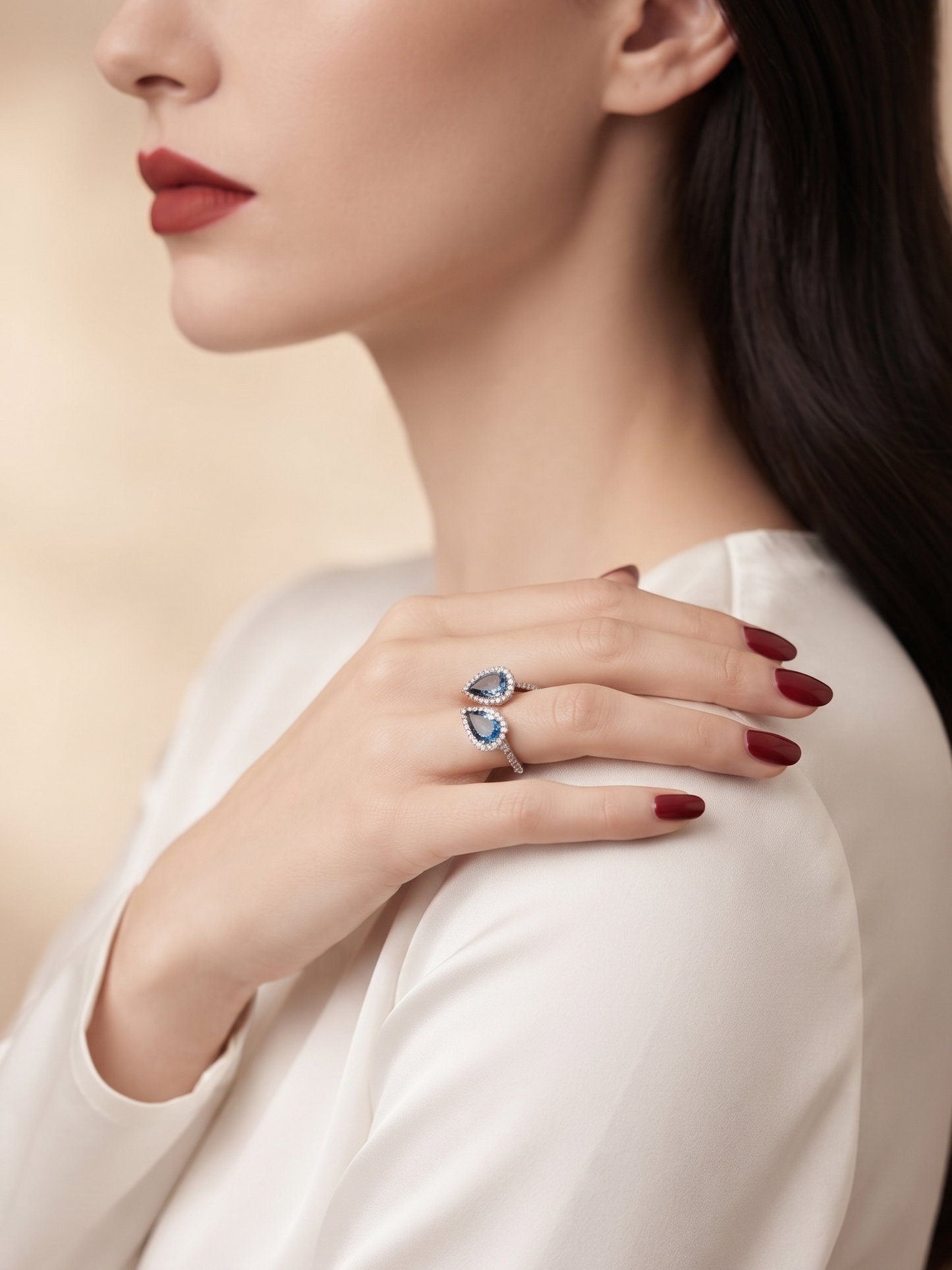 A close-up lifestyle shot of a woman wearing a silver ring with two blue teardrop-shaped gemstones on her finger, wearing a white satin blouse against a soft beige background.