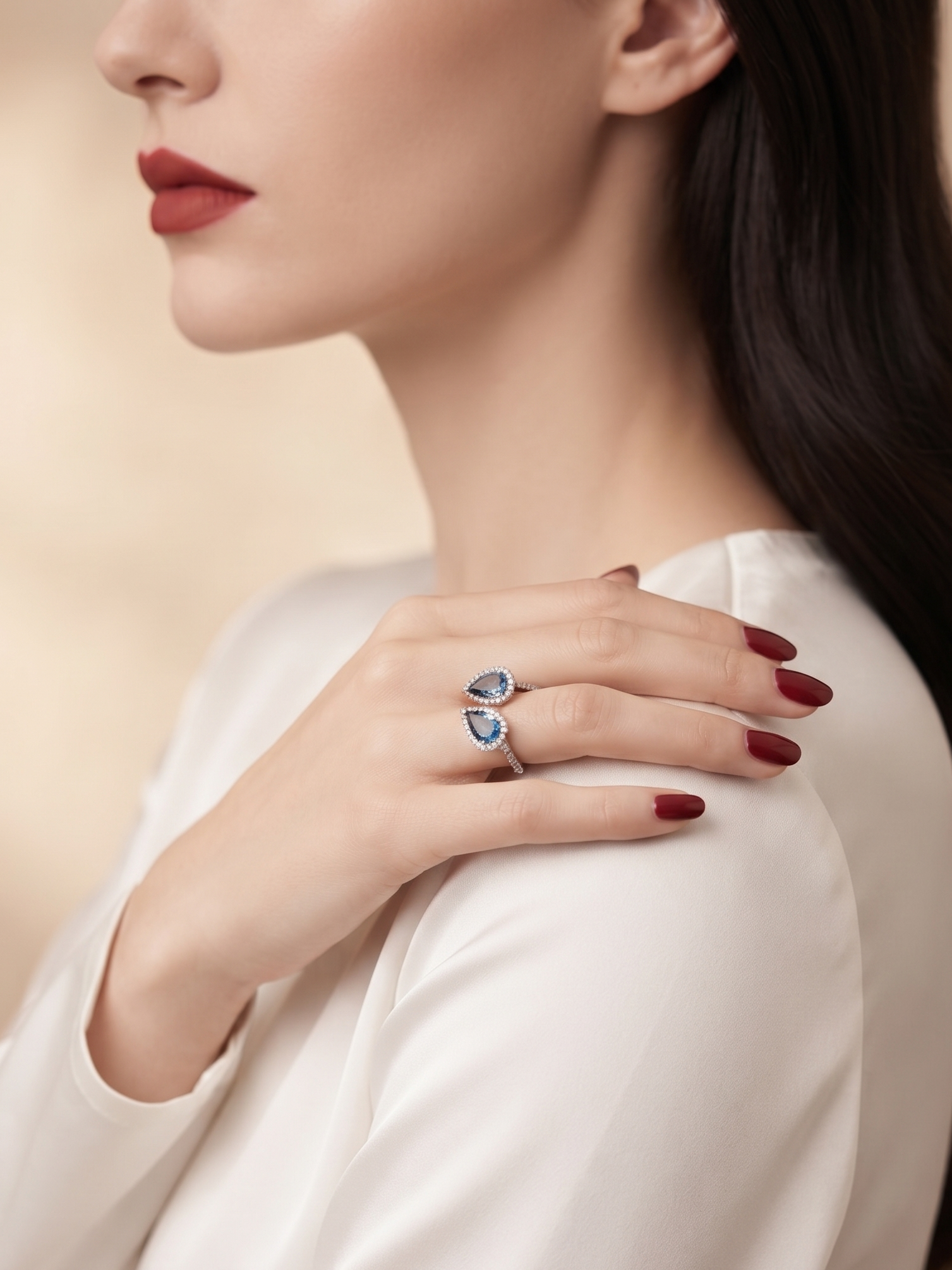 A close-up lifestyle shot of a woman wearing a silver ring with two blue teardrop-shaped gemstones on her finger, wearing a white satin blouse against a soft beige background.
