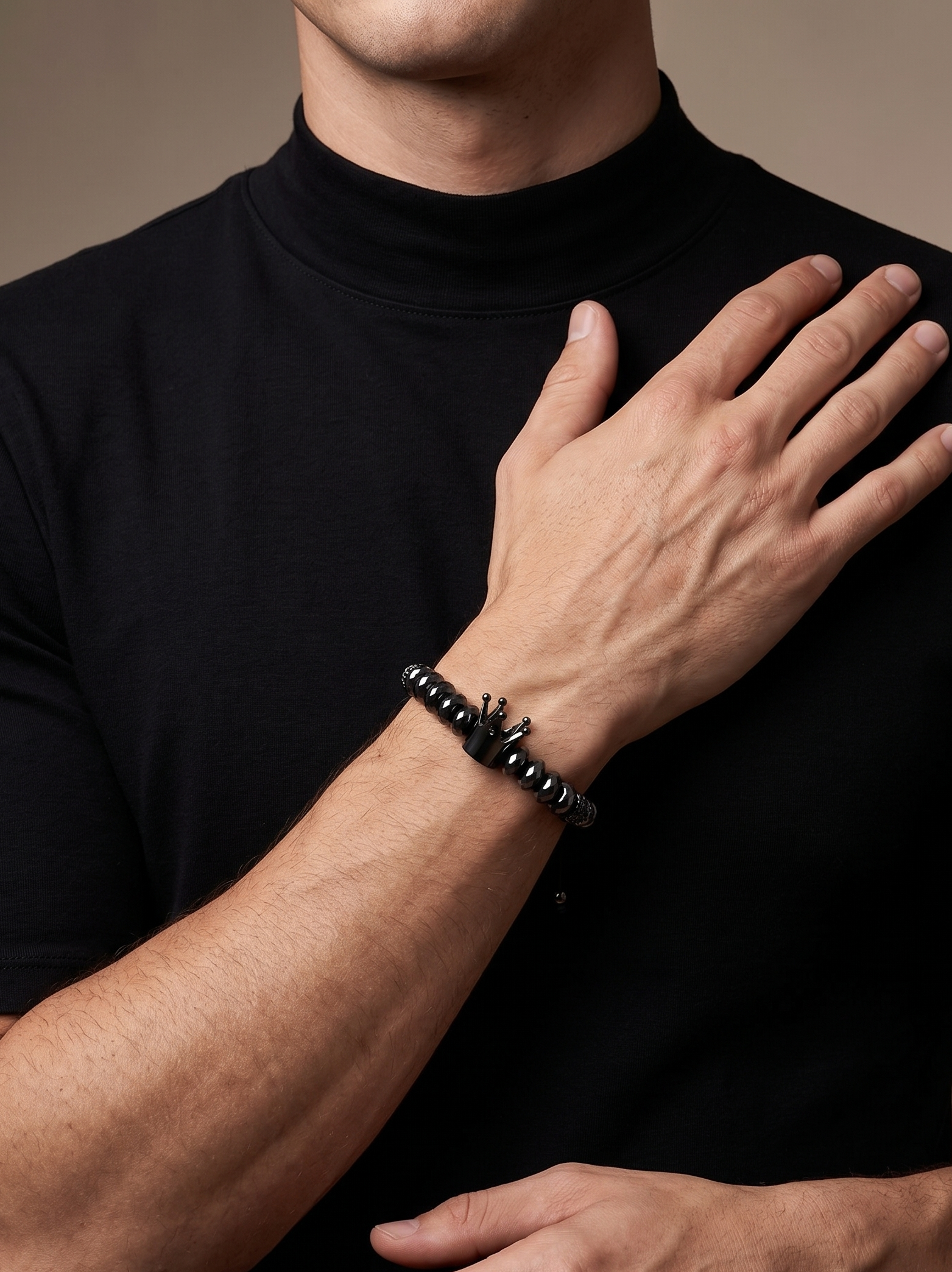 A close-up of a man wearing a black beaded bracelet with a small silver-toned pendant on his left wrist, wearing a black turtleneck against a warm beige background.