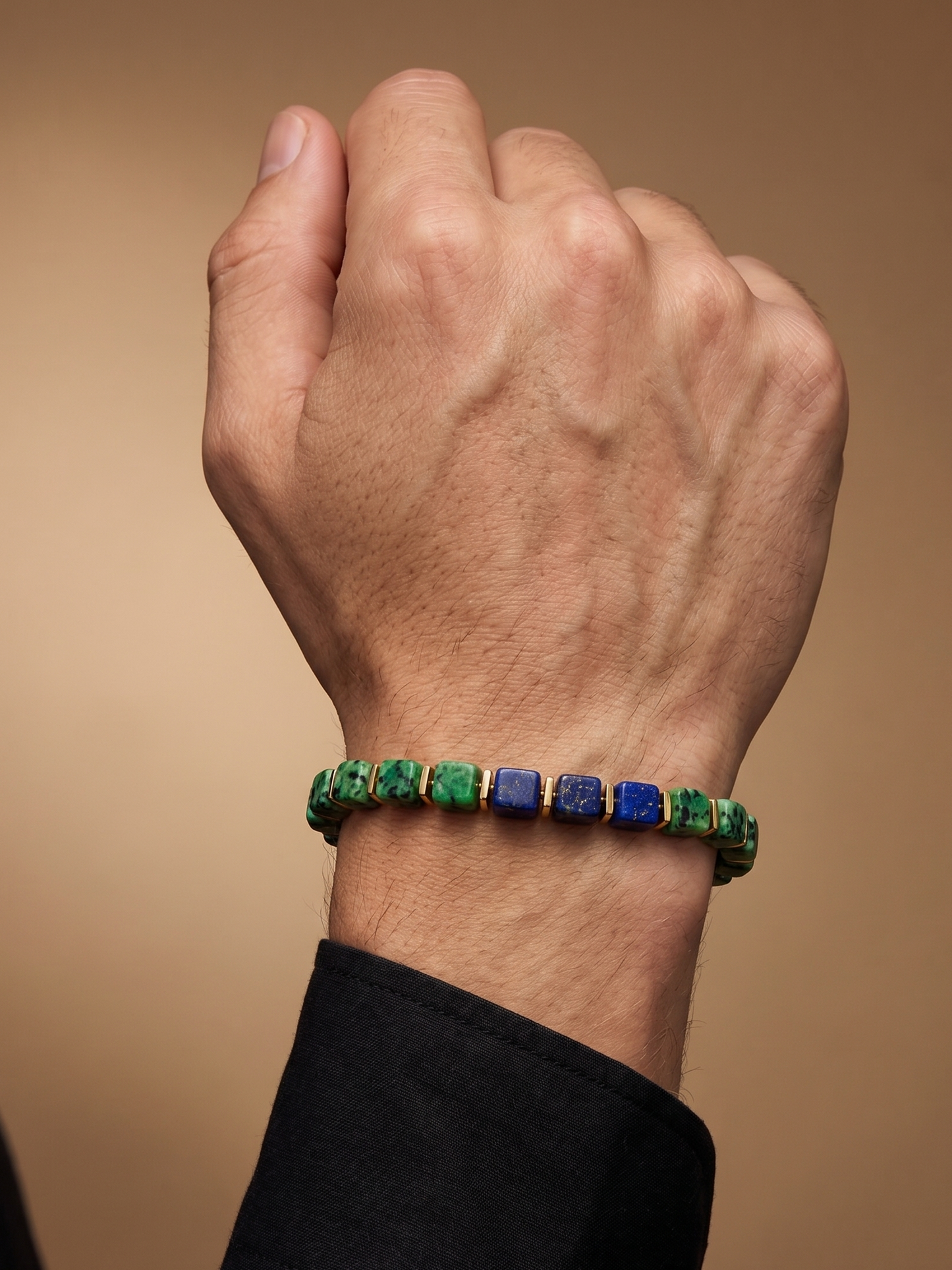 A close-up of a person's wrist wearing a beaded bracelet with alternating green and blue square beads, paired with a black dress shirt cuff against a warm brown background.