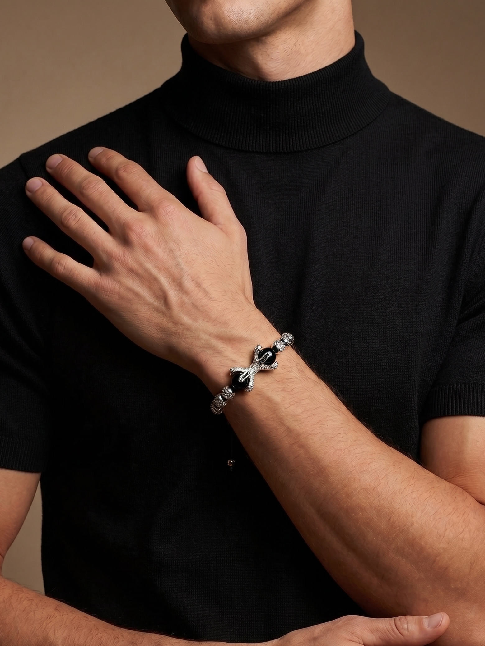 A close-up of a man wearing a black turtleneck sweater and a silver-toned bracelet with black beads and a bow detail on his wrist, set against a warm brown studio background.