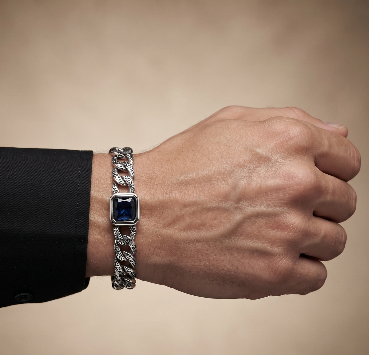 A close-up of a man's hand and wrist wearing a silver-toned chain bracelet with a square dark blue gemstone centerpiece, paired with a black dress shirt cuff.
