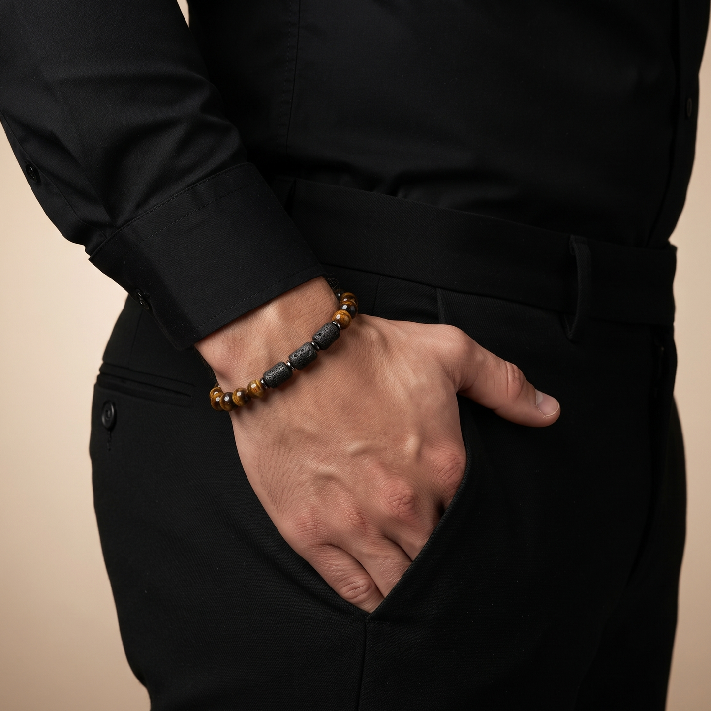 A close-up of a man wearing a black long-sleeve shirt and black trousers, with his hand in his pocket, showcasing a beaded bracelet on his wrist. The bracelet features alternating translucent amber-brown beads and dark gray beads.