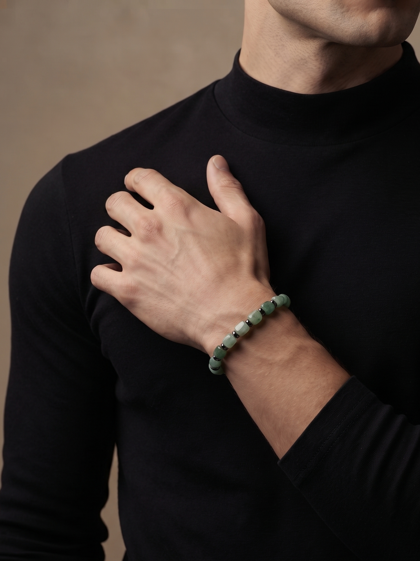 A close-up of a man wearing a black long-sleeve turtleneck, showcasing a green beaded bracelet on his wrist against a warm, neutral background.