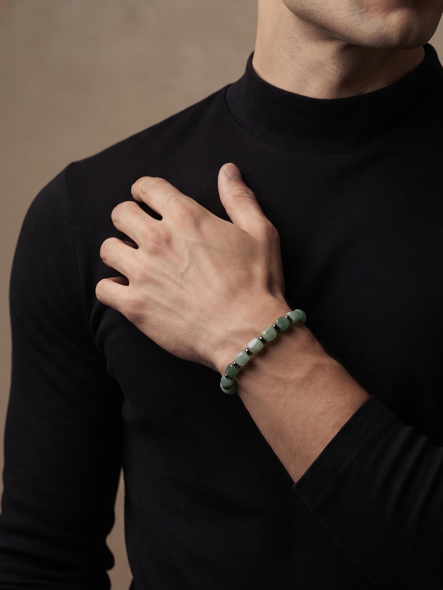 A close-up of a man wearing a black long-sleeve turtleneck, showcasing a green beaded bracelet on his wrist against a warm, neutral background.