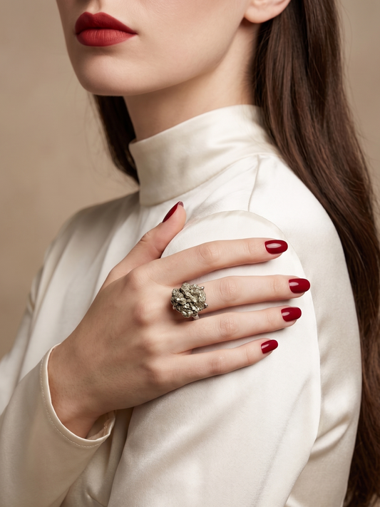 A close-up of a woman wearing a large, ornate silver ring with a floral or leafy design on her ring finger, paired with a cream satin mock-neck top and dark red nail polish against a warm beige background.