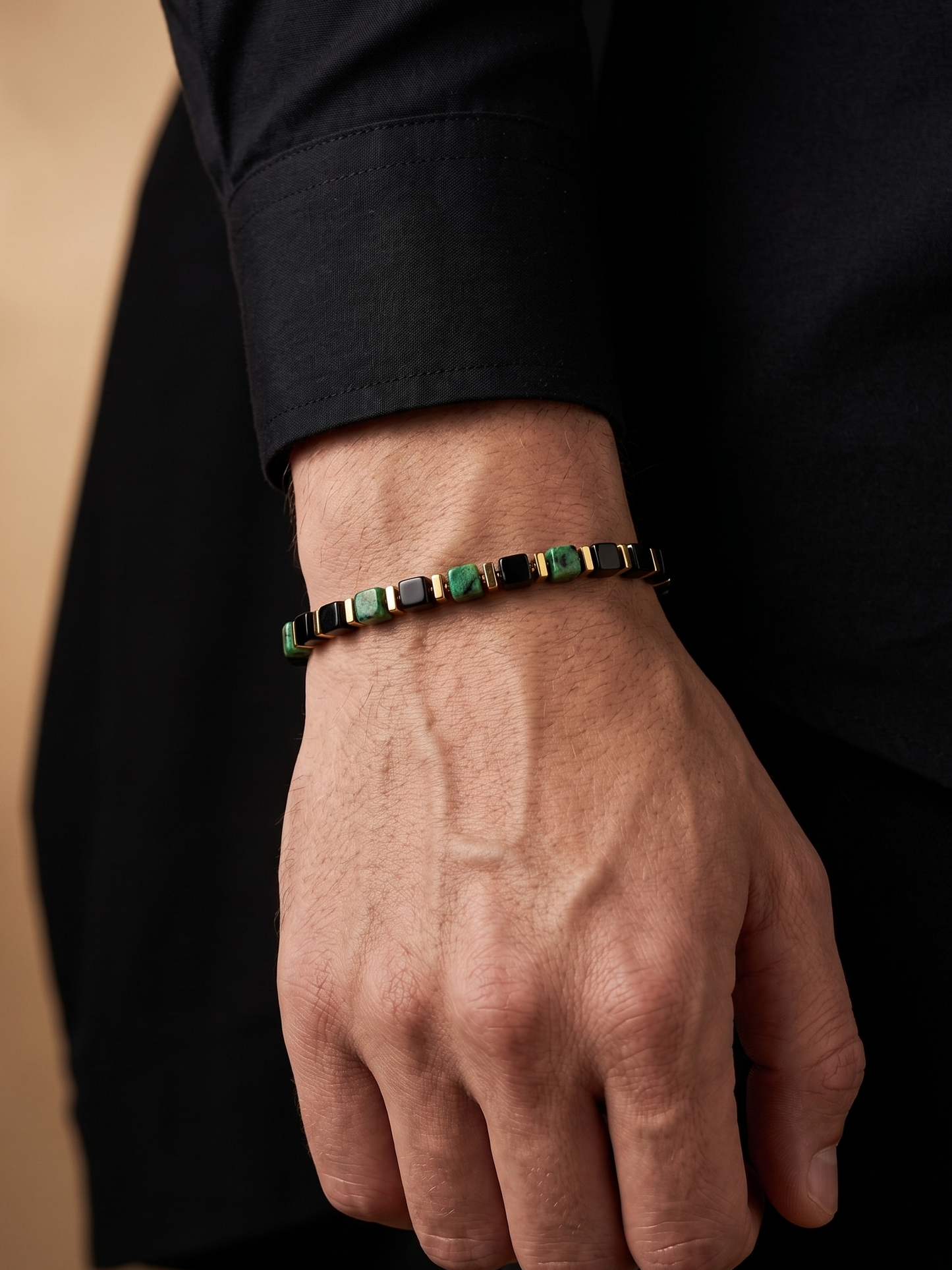 A close-up of a man's hand and wrist wearing a slim beaded bracelet made of alternating dark green and black square beads, styled with a black dress shirt.