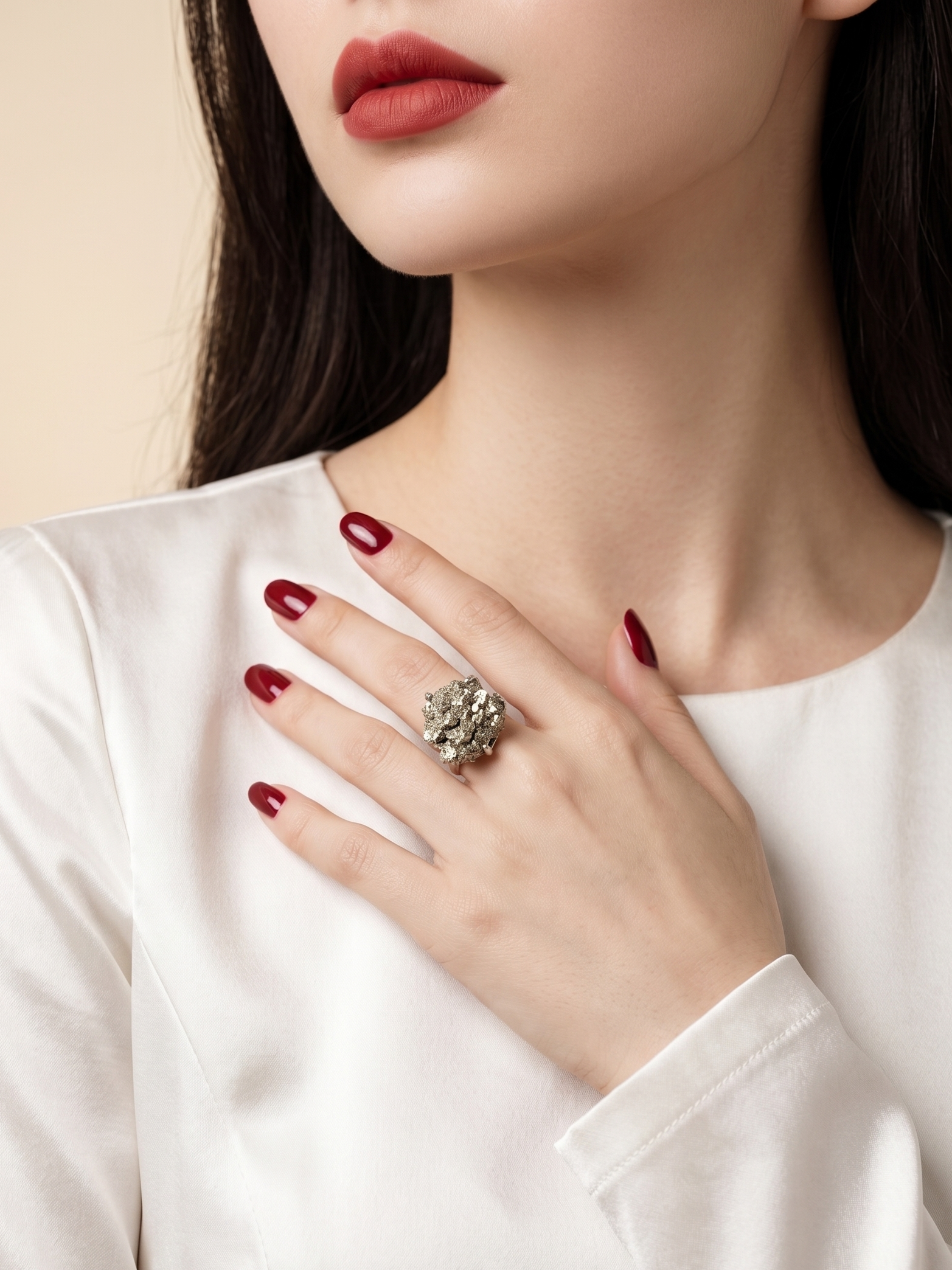 A close-up lifestyle shot of a model wearing a large, ornate silver ring with a floral cluster of clear stones on her hand, styled with dark red nail polish and a white satin top against a soft beige background.