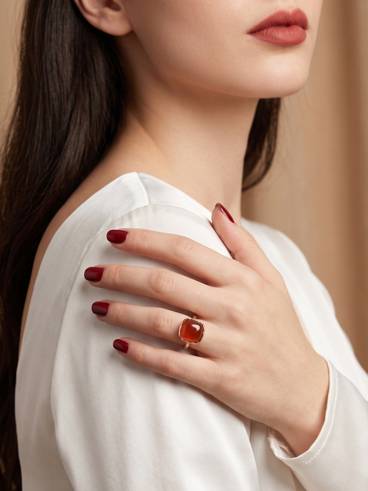 A close-up lifestyle shot of a woman wearing a gold ring with a large, polished orange cabochon gemstone on her ring finger, paired with a white satin blouse and dark red manicured nails.