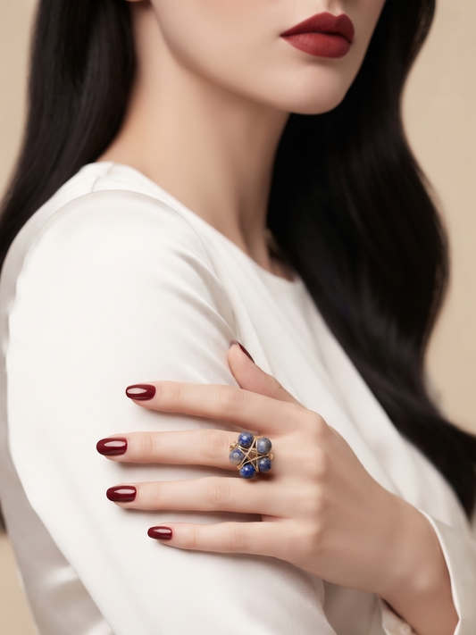 A close-up lifestyle shot of a model wearing a distinctive flower-shaped ring with blue and gold details on her finger, dressed in a white satin top against a warm beige background.