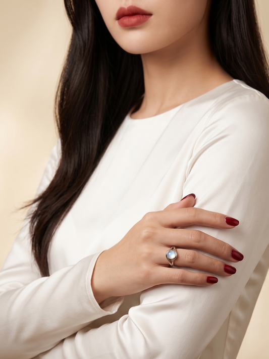 A close-up of a woman wearing a white long-sleeve top and a silver ring with a light blue stone, shown from the chest up against a soft beige background.