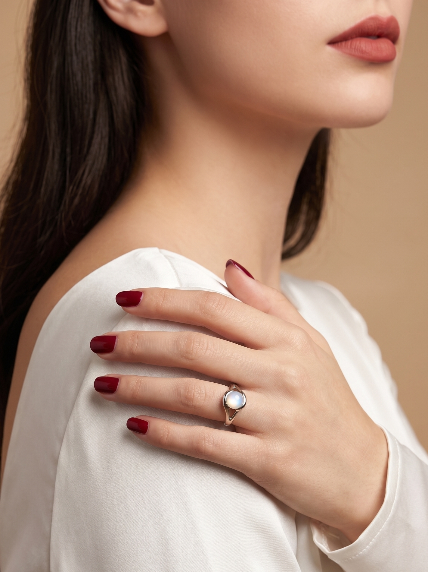 A close-up lifestyle shot of a woman wearing a silver ring with a round white stone, resting her hand on her shoulder. She is wearing a white top and has dark red manicured nails, set against a solid warm beige background.