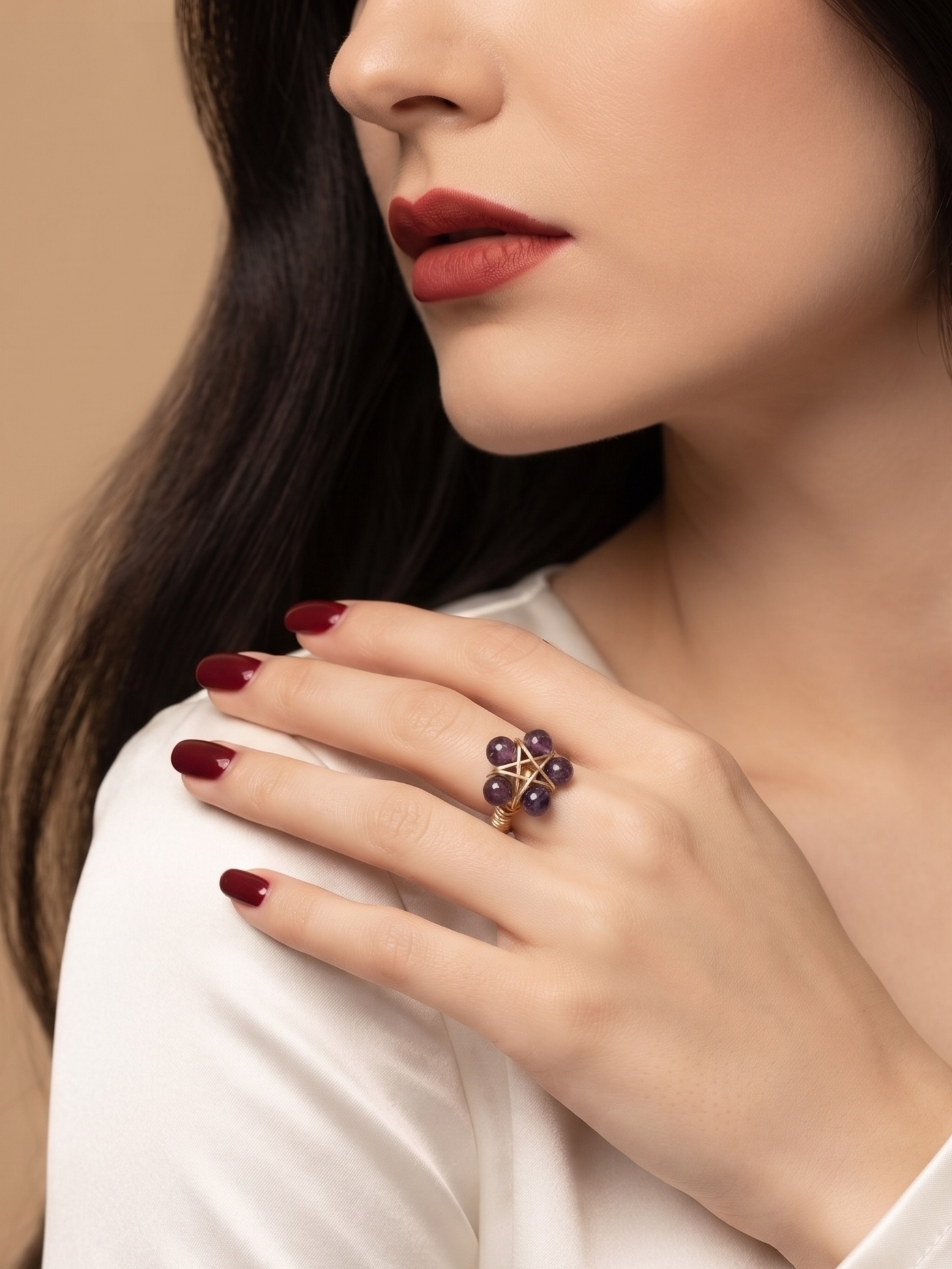 A close-up lifestyle shot of a woman wearing a gold ring with four dark purple gemstone clusters, styled with dark red lipstick and manicured nails on a white satin blouse against a warm beige background.