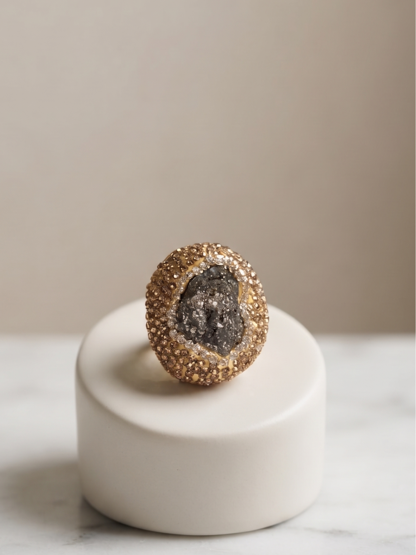 A gold ring with a textured, geode-like dark gray center, displayed on a small white cylindrical pedestal over a white marble surface against a neutral beige background.