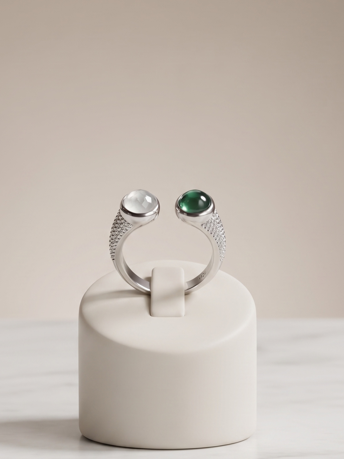 A silver ring displayed on a small cream-colored stand, featuring a clear oval gemstone on one side and a green oval gemstone on the other, set against a neutral beige background and white marble surface.