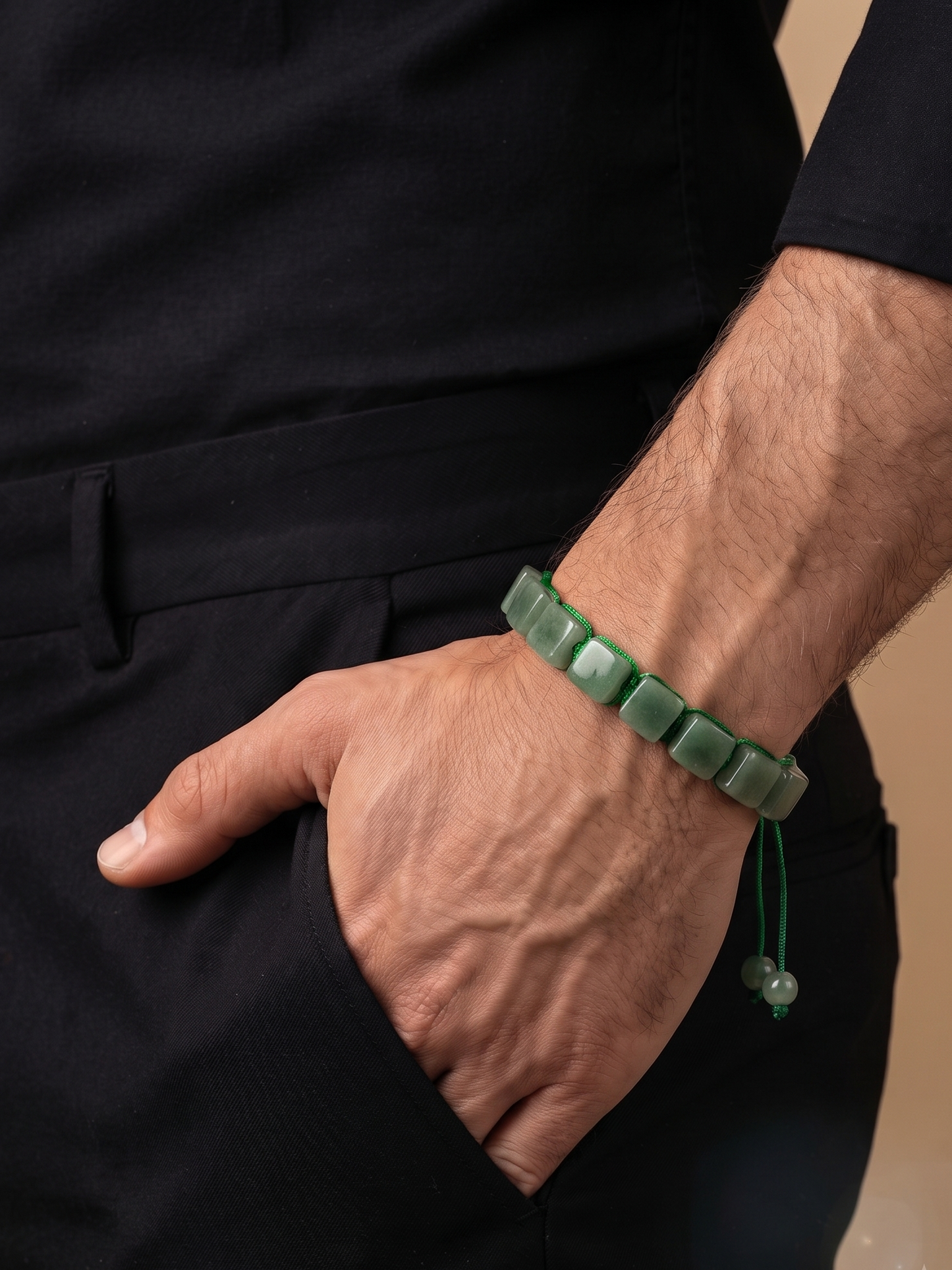 A close-up of a man's hand in his pocket wearing a green square-beaded bracelet with a matching cord tassel, paired with a black shirt and trousers.