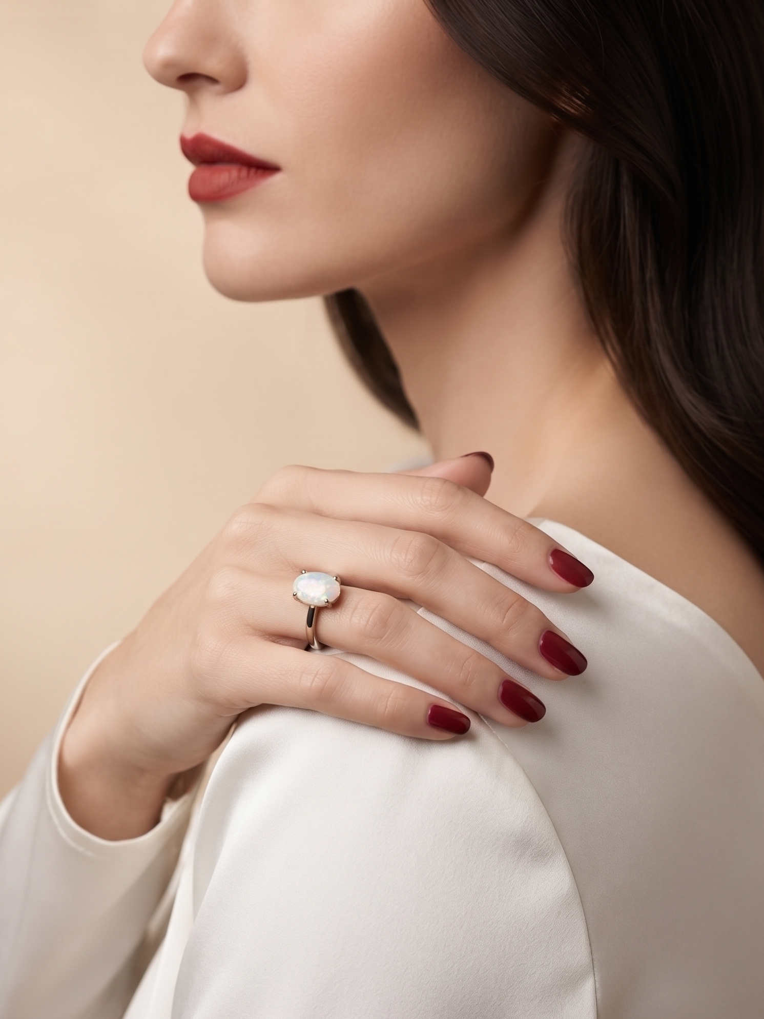 A close-up lifestyle shot of a woman wearing a silver ring with a large white oval gemstone, styled with dark red nail polish and a white off-the-shoulder top against a warm beige background.