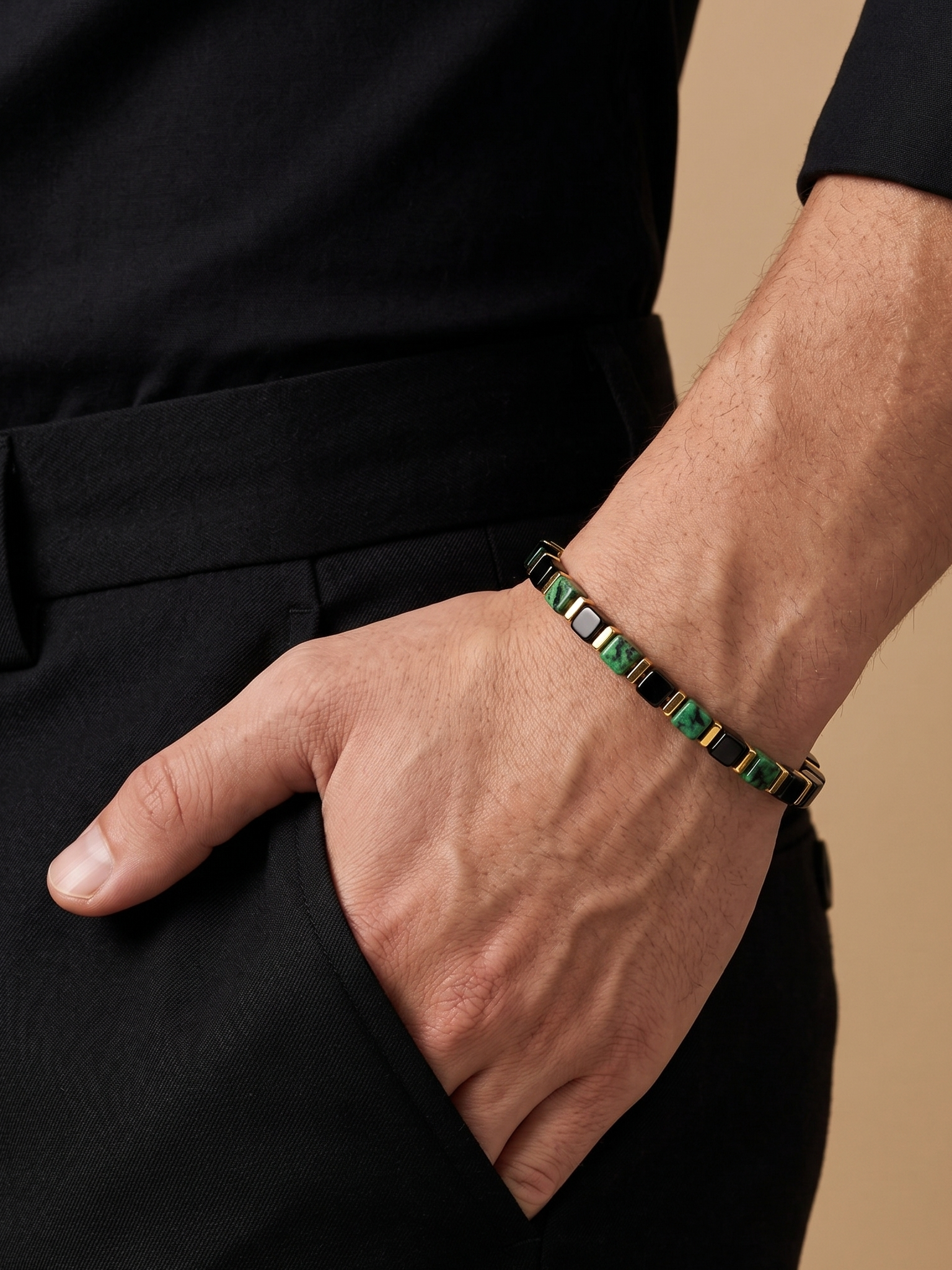 A close-up of a man's hand in his pocket wearing a green and black beaded bracelet with gold-toned metal accents, paired with a black shirt and trousers against a warm beige background.