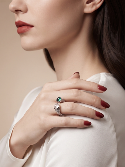 A close-up lifestyle shot of a woman wearing two silver rings on her fingers, one with a green gemstone and the other with a clear gemstone, while wearing a white satin top against a warm beige background.