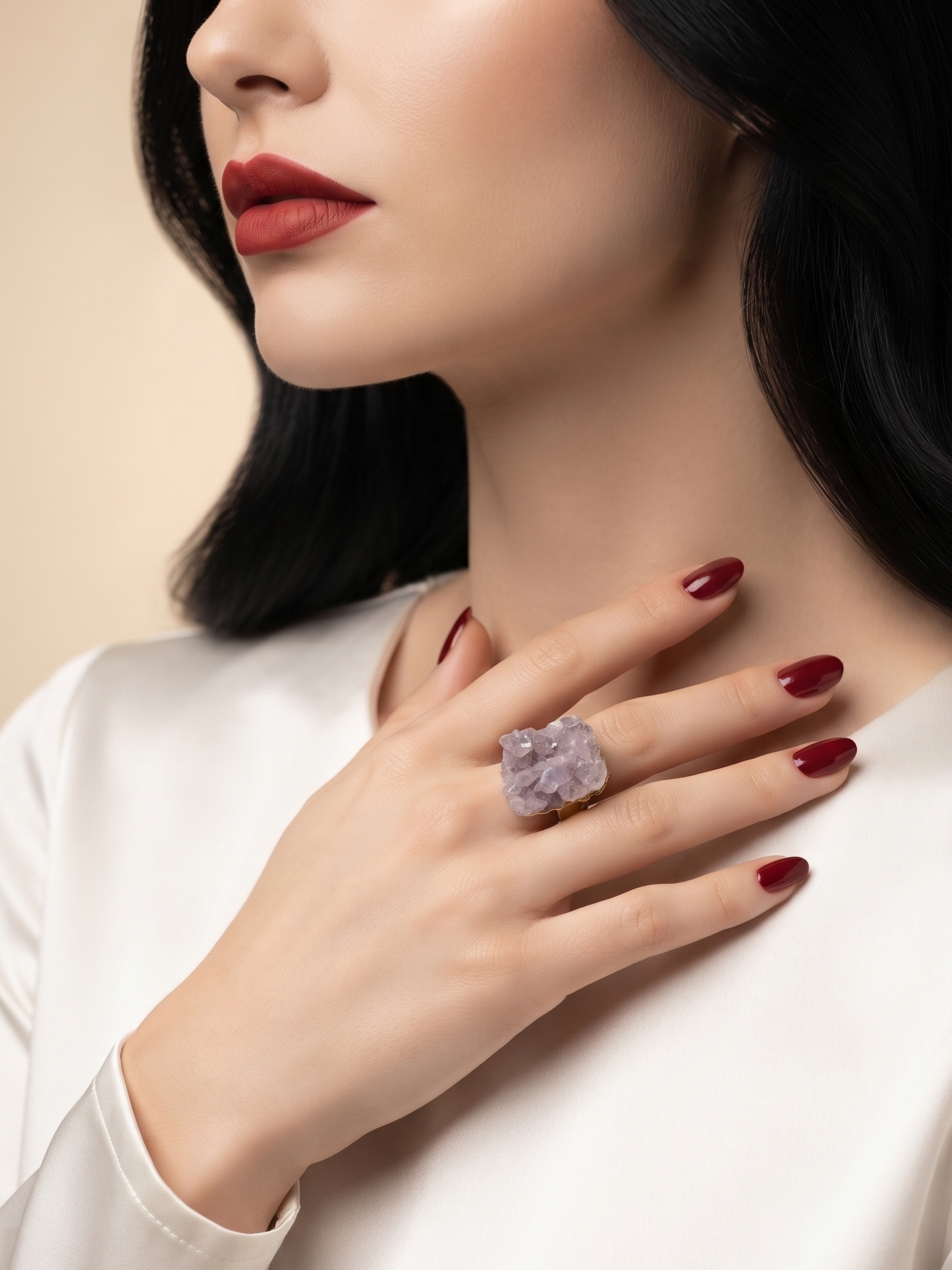 A close-up lifestyle shot of a woman wearing a large, chunky lavender crystal ring on her finger, dressed in a white satin top against a soft beige background.