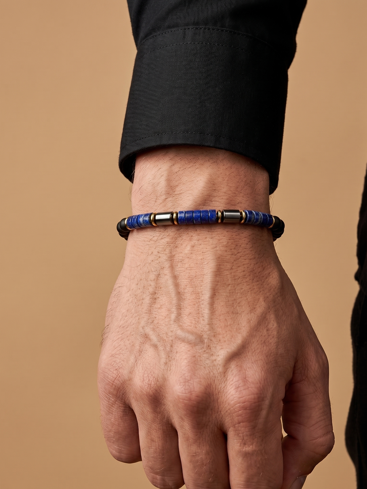A close-up of a person's hand wearing a beaded bracelet with blue, black, and metallic gold beads, set against a solid tan background.