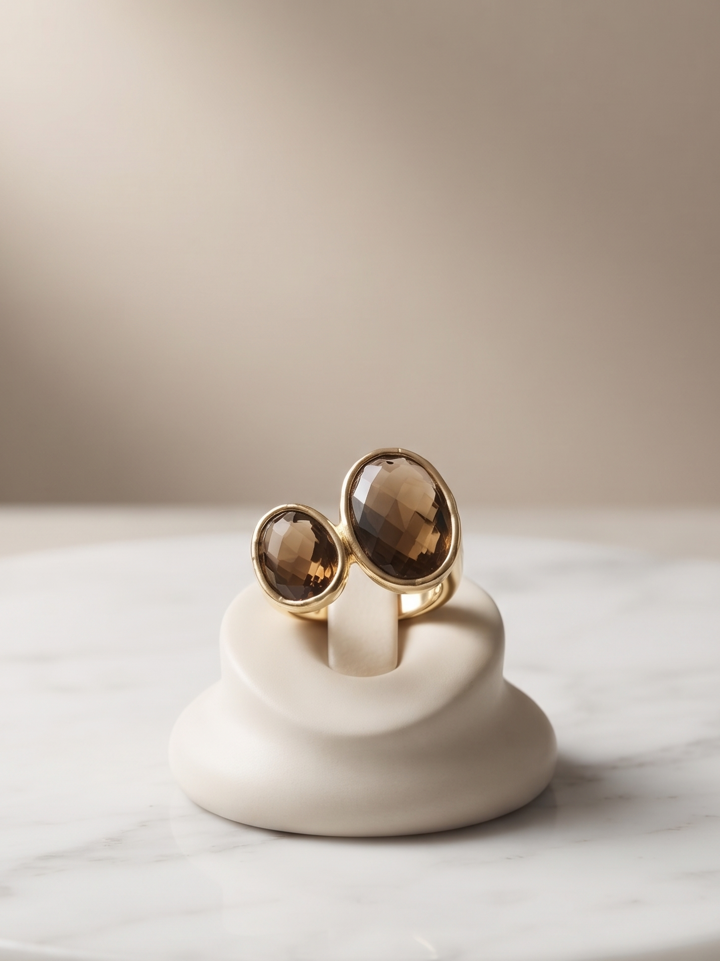 Two gold-toned rings with oval smoky brown gemstones displayed on a small white ceramic stand, set on a round white marble slab against a soft beige background.