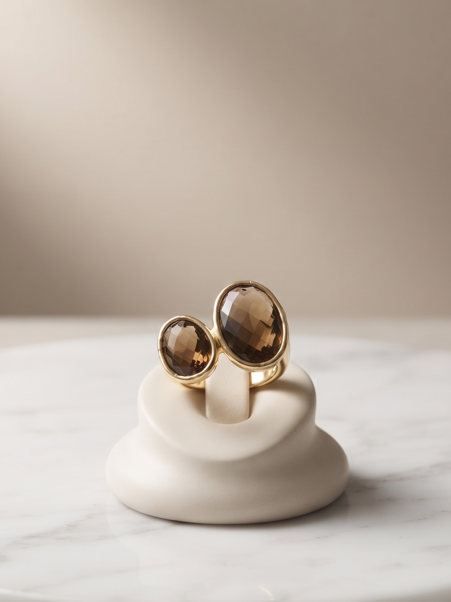 Two gold-toned rings with oval smoky brown gemstones displayed on a small white ceramic stand, set on a round white marble slab against a soft beige background.