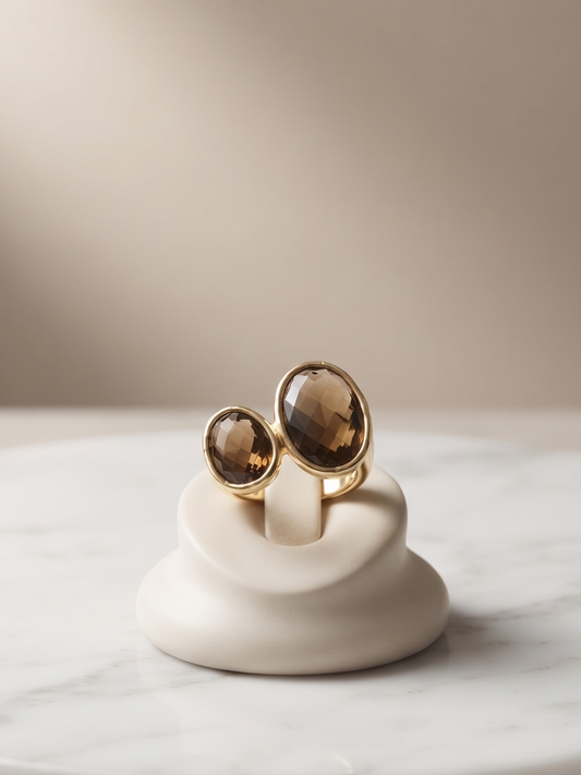 Two gold-toned rings with oval smoky brown gemstones displayed on a small white ceramic stand, set on a round white marble slab against a soft beige background.