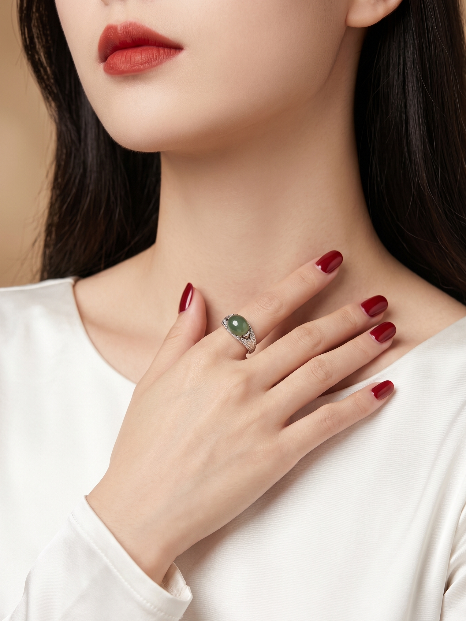 A close-up of a woman wearing a silver ring with a green oval stone, with her hand resting on her chest. She is wearing a white long-sleeve top and has dark red nail polish.