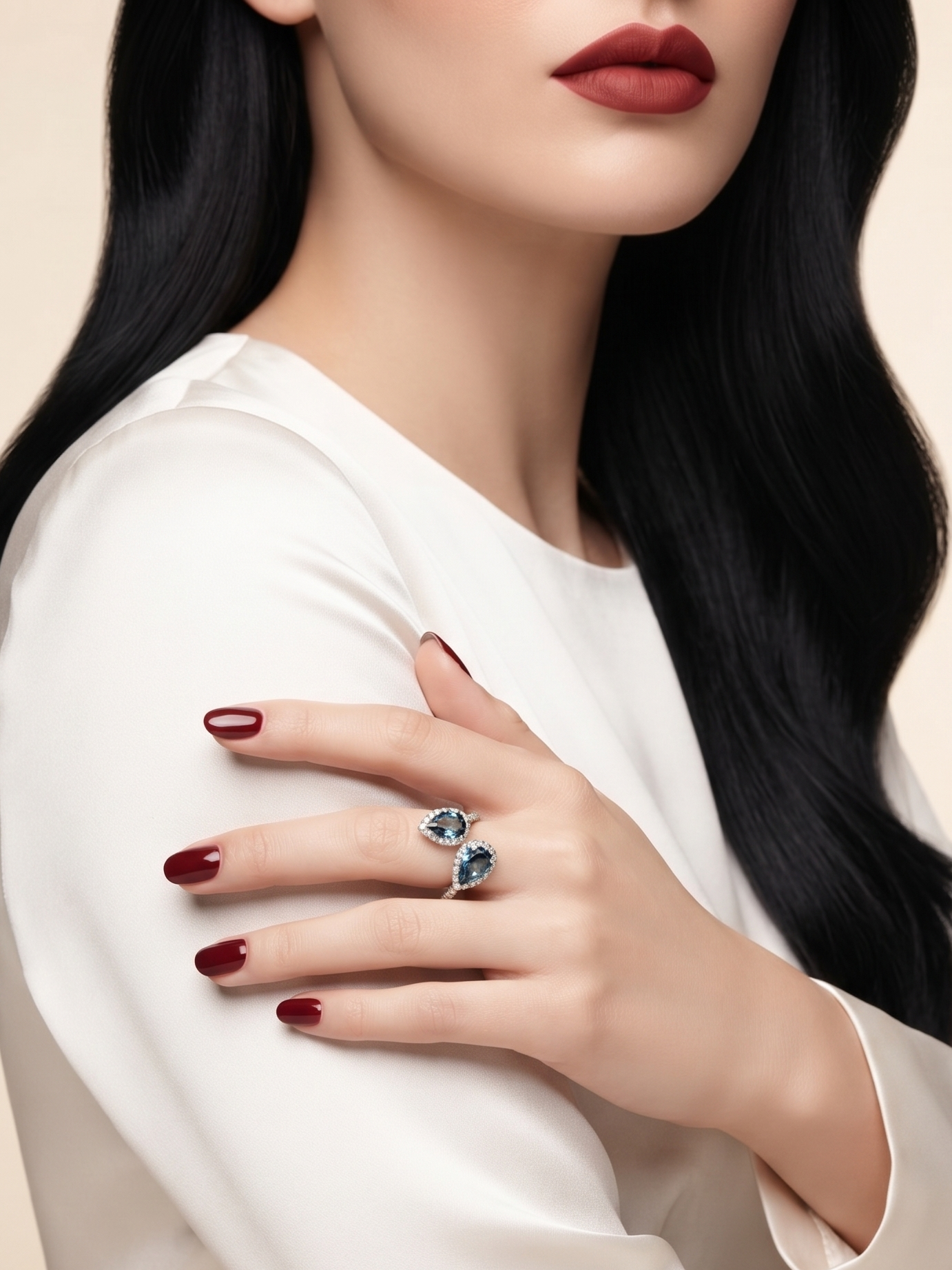 A close-up of a woman wearing a white satin long-sleeve top, showcasing a silver ring with a marquise-cut blue gemstone on her hand with dark red nail polish against a warm beige background.