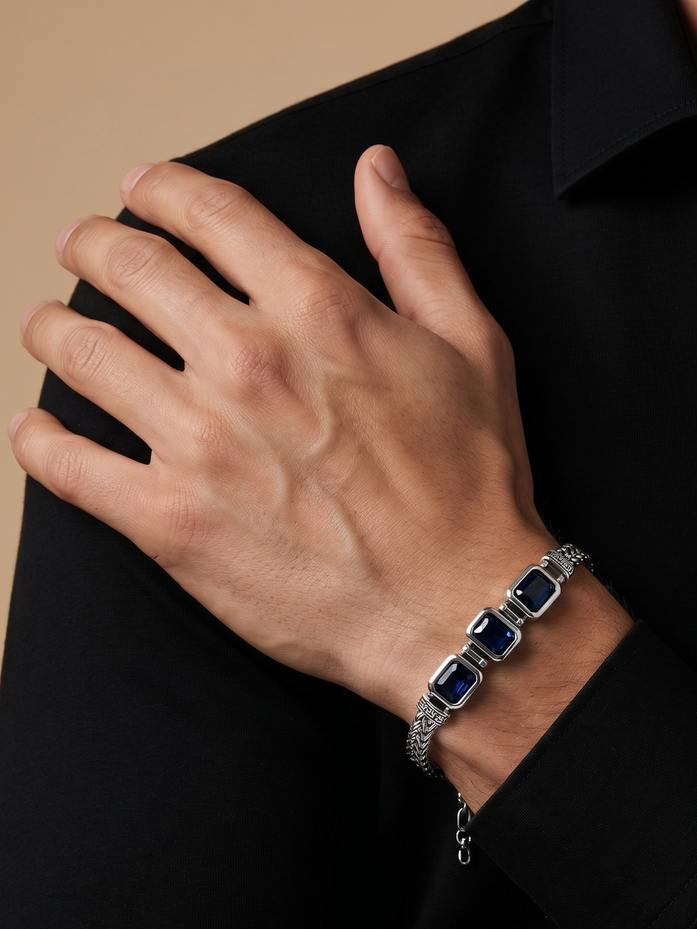 A close-up of a man wearing a silver-toned bracelet with rectangular dark blue stones, shown on his wrist against a black shirt and warm beige background.