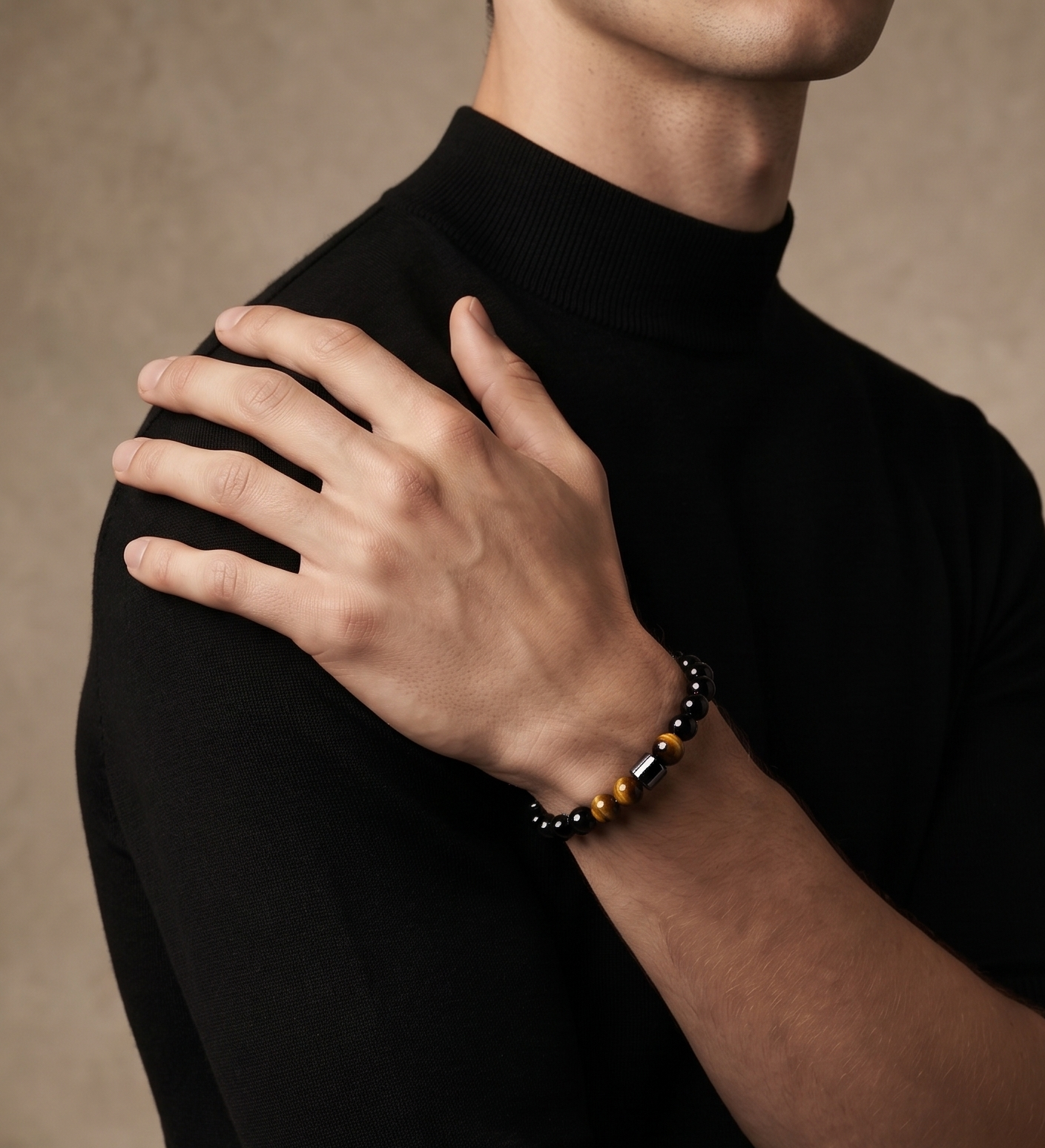A close-up of a man wearing a black long-sleeve mock neck t-shirt and a beaded bracelet on his wrist, with his hand resting on his shoulder, set against a textured warm brown background.